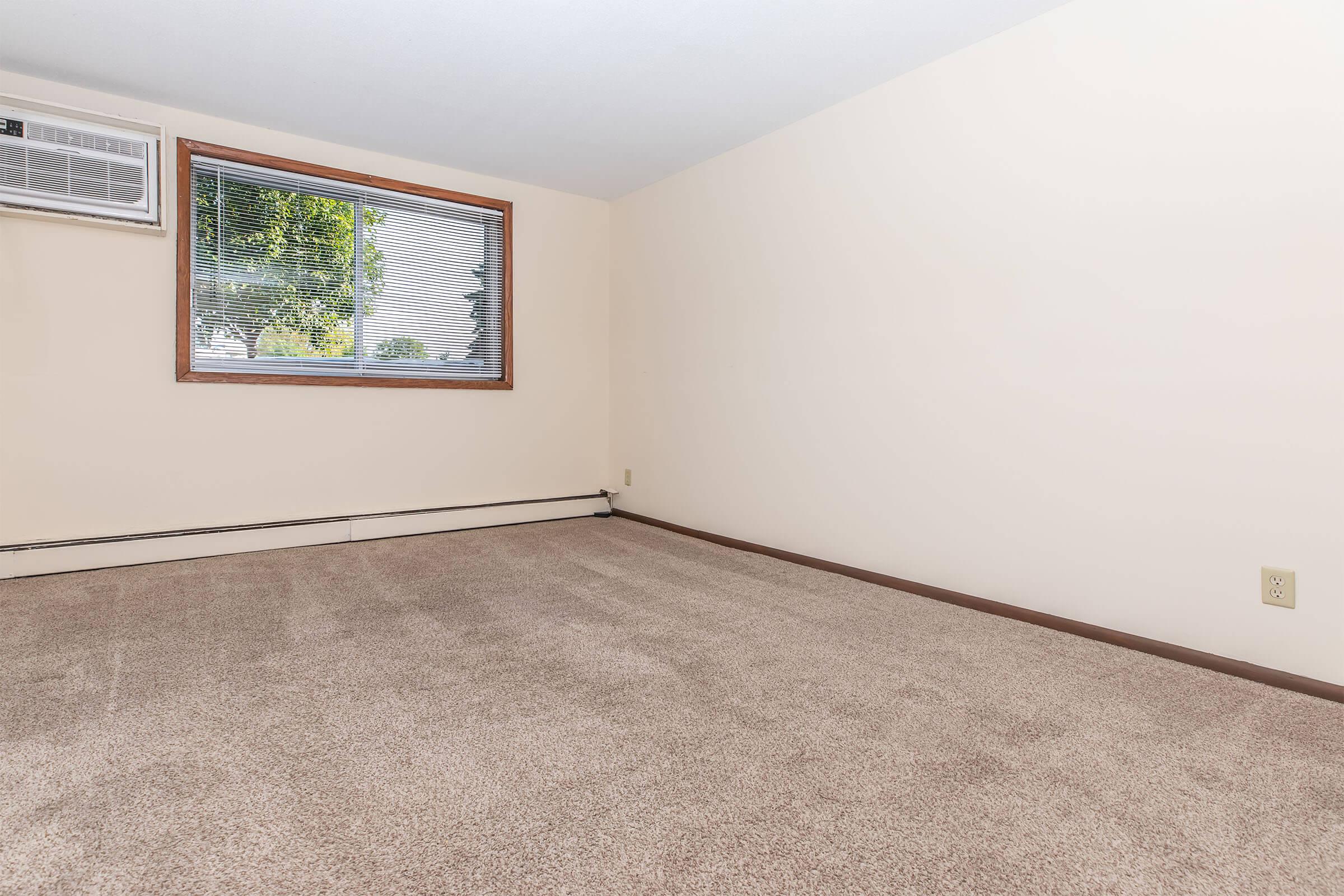 A vacant room with beige walls and light brown carpeting. There is a window with blinds on the left side, allowing natural light to enter. An air conditioning unit is installed beneath the window, and the room has a simple, uncluttered appearance with no furniture or decorations.