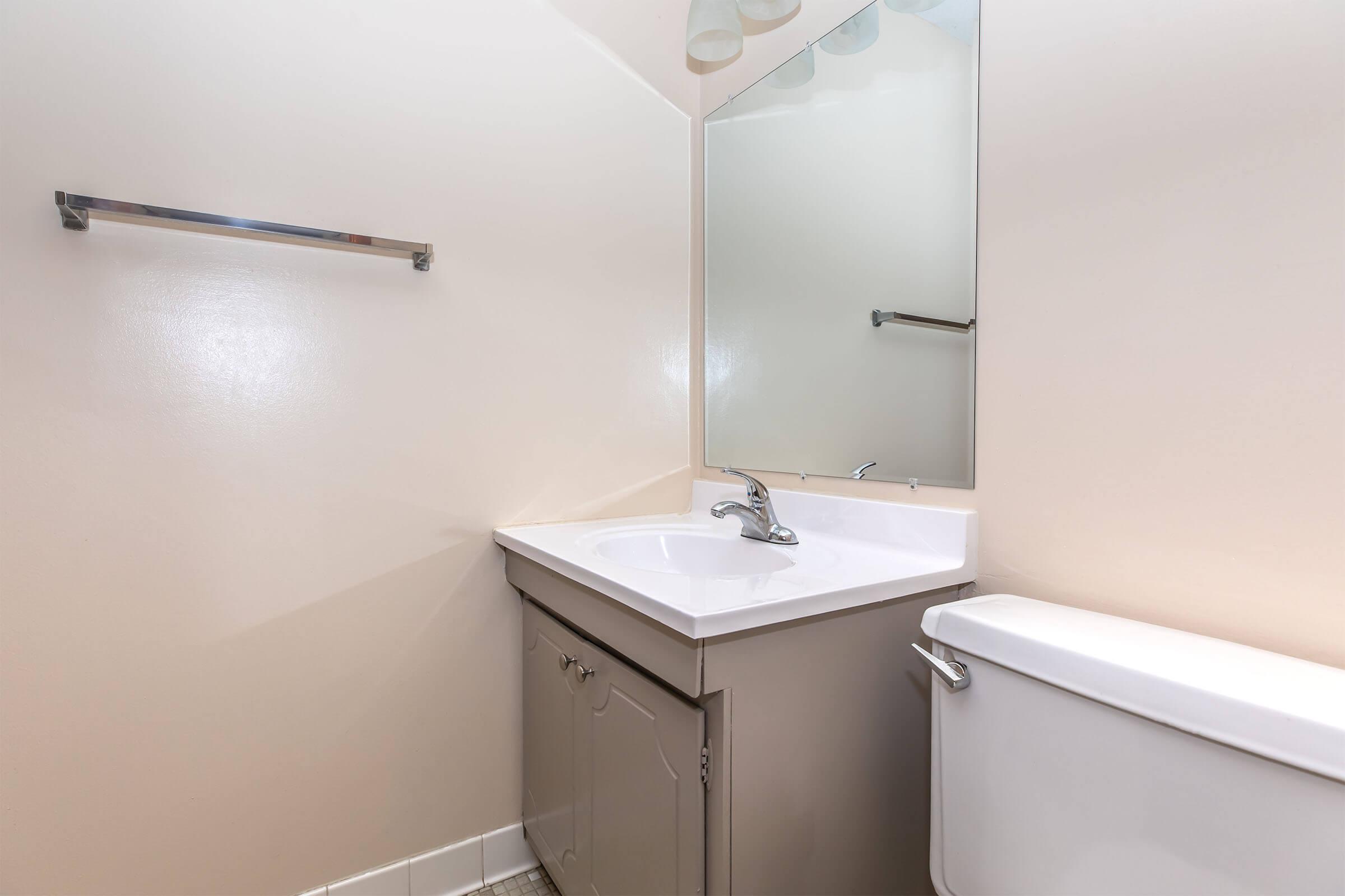 A small bathroom featuring a white sink with a chrome faucet, a light-colored countertop, a mirror above the sink, a toilet on the right, and a towel rack on the wall. The walls are painted in a neutral tone, creating a simple and clean atmosphere.