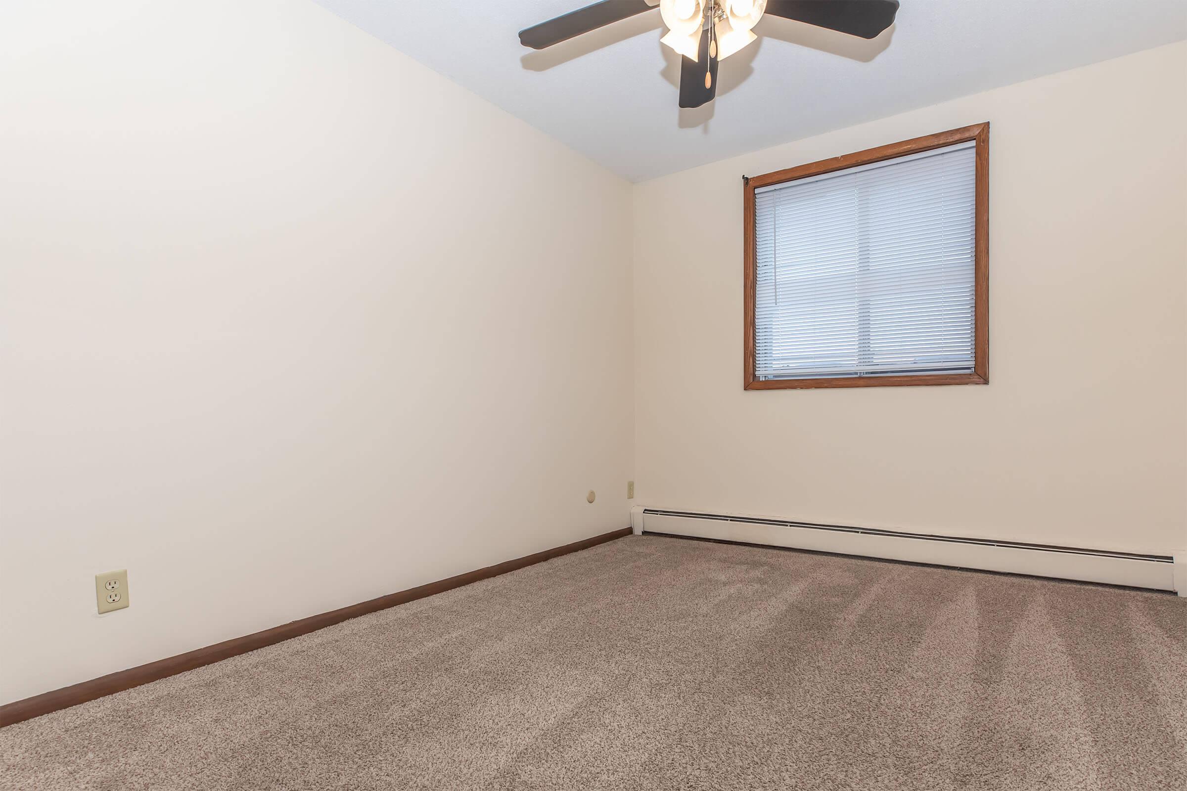 A blank room with light beige walls and a window covered by blinds. The floor is carpeted in a light color, and there is a ceiling fan with three blades. A baseboard heater is visible along the wall. The room appears empty, providing a spacious feel.