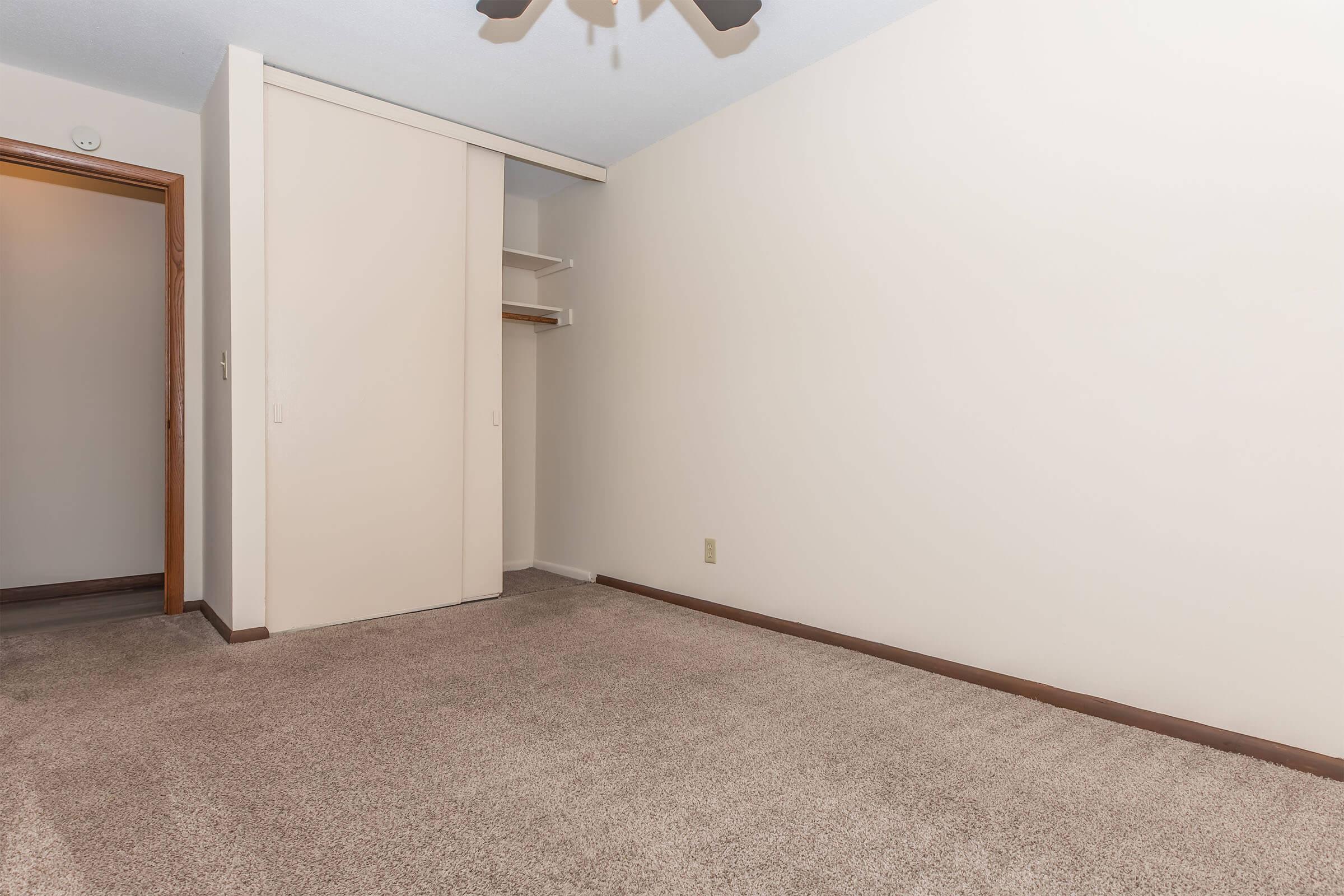 Empty room with light beige walls and carpeted flooring. There is a sliding closet door on the left side and a ceiling fan. A doorway is visible on the left, leading to another area. The space appears clean and unoccupied, suitable for various uses.