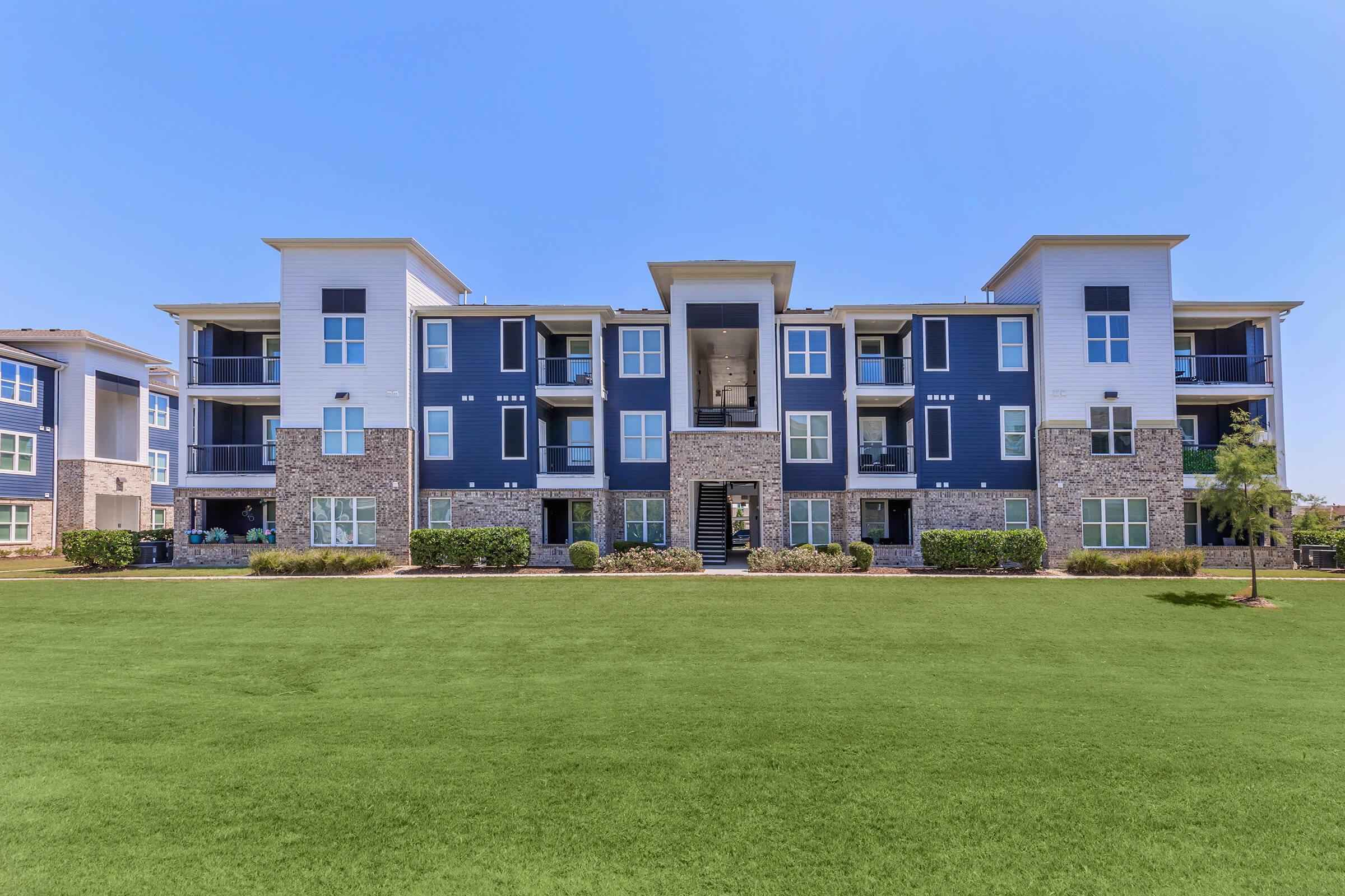 A modern apartment building featuring a mix of stone and siding exteriors. The structure is three stories tall with multiple balconies and large windows. A well-maintained lawn surrounds the building, providing a green space in front. Clear blue sky adds to the bright and welcoming atmosphere.