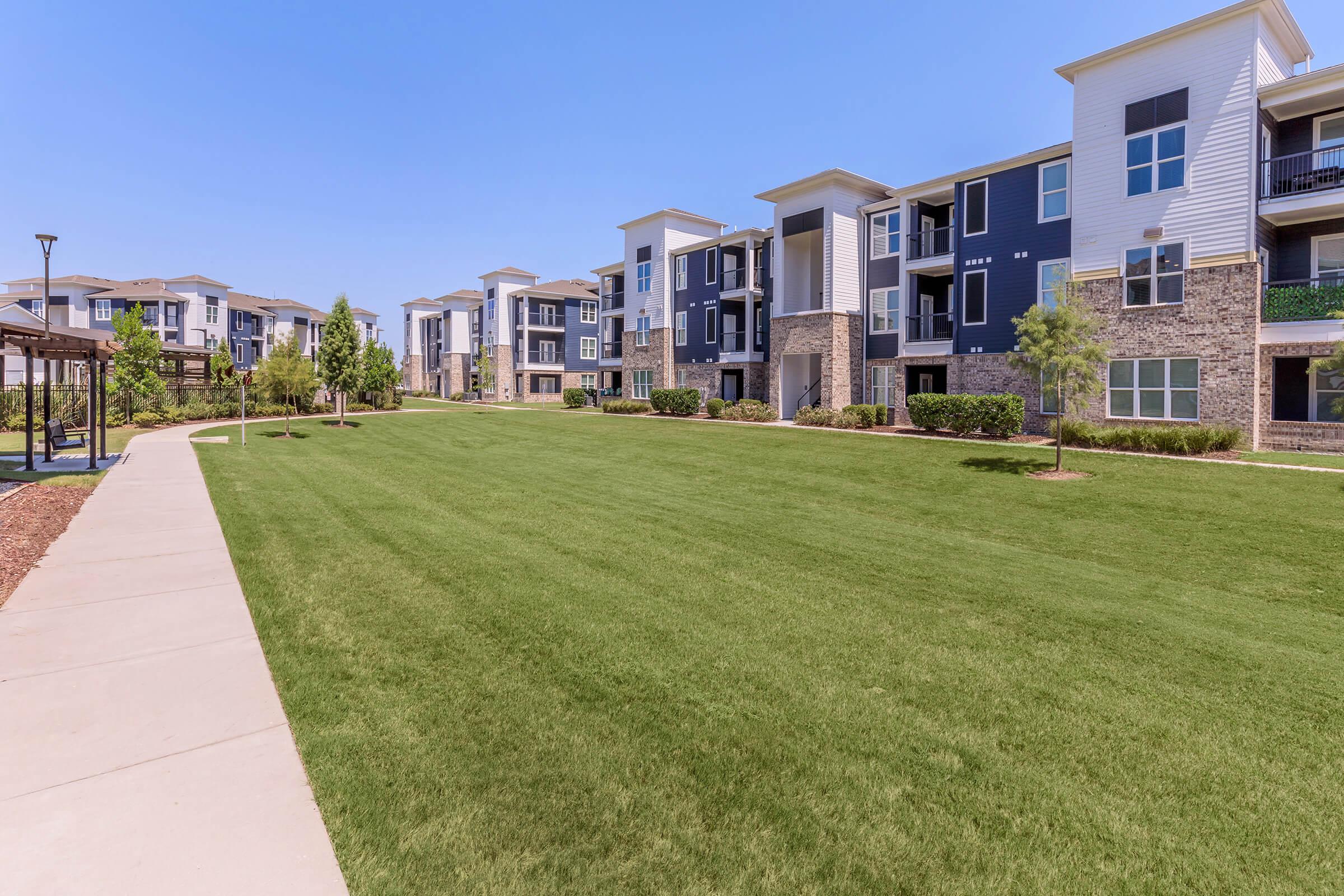 A well-maintained apartment complex with multiple modern buildings. The scene features green lawns, neatly trimmed bushes, and a paved walkway. Clear blue skies and ample sunlight create a bright and inviting atmosphere in the outdoor living space.