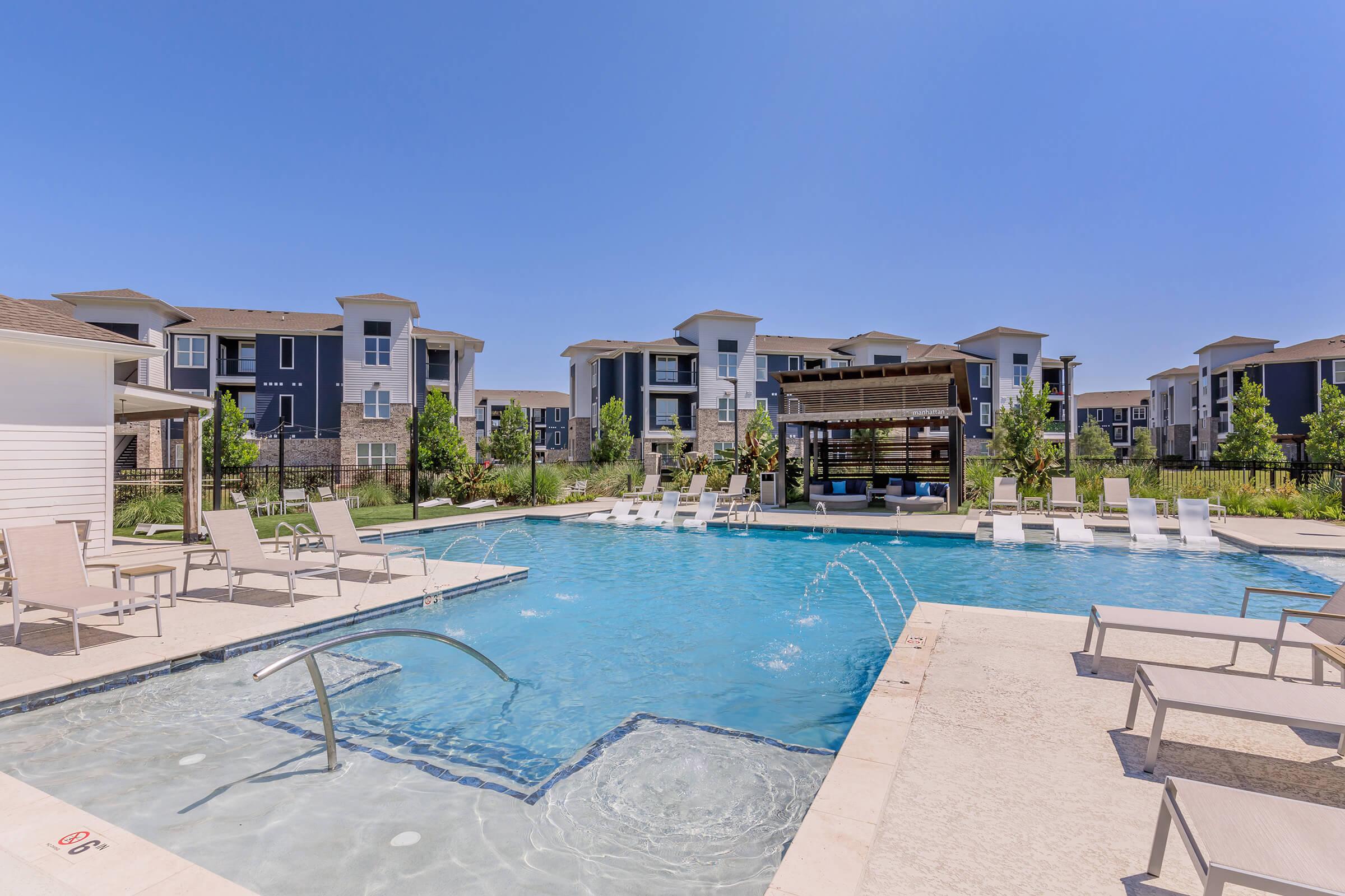 A clear blue swimming pool surrounded by lounge chairs, with a wooden pergola nearby. Modern apartment buildings are visible in the background under a bright blue sky. The area is well-maintained with greenery and features a spacious deck for relaxation.