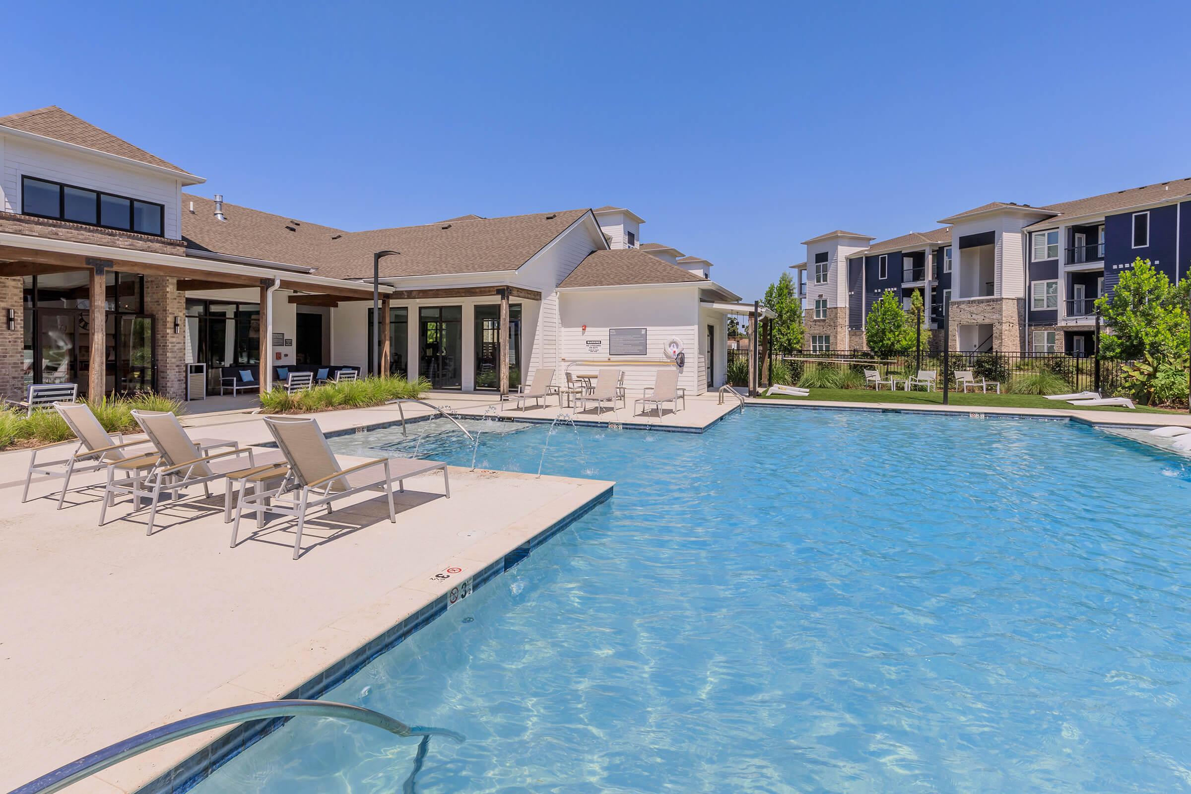 Swimming pool area at a residential complex, featuring clear blue water, lounge chairs, and modern buildings in the background. Sunlight brightens the scene, creating a relaxing atmosphere. Plant beds add greenery around the pool area, inviting residents to enjoy the space.