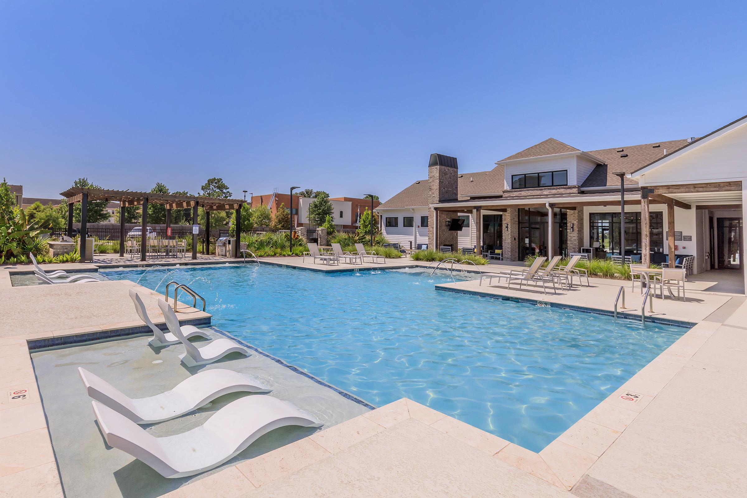 A spacious outdoor pool area featuring a large, clear blue swimming pool with lounge chairs, an adjacent hot tub, and a shaded pergola. Surrounding the pool are well-maintained landscaping and modern buildings, creating a relaxing atmosphere on a sunny day.