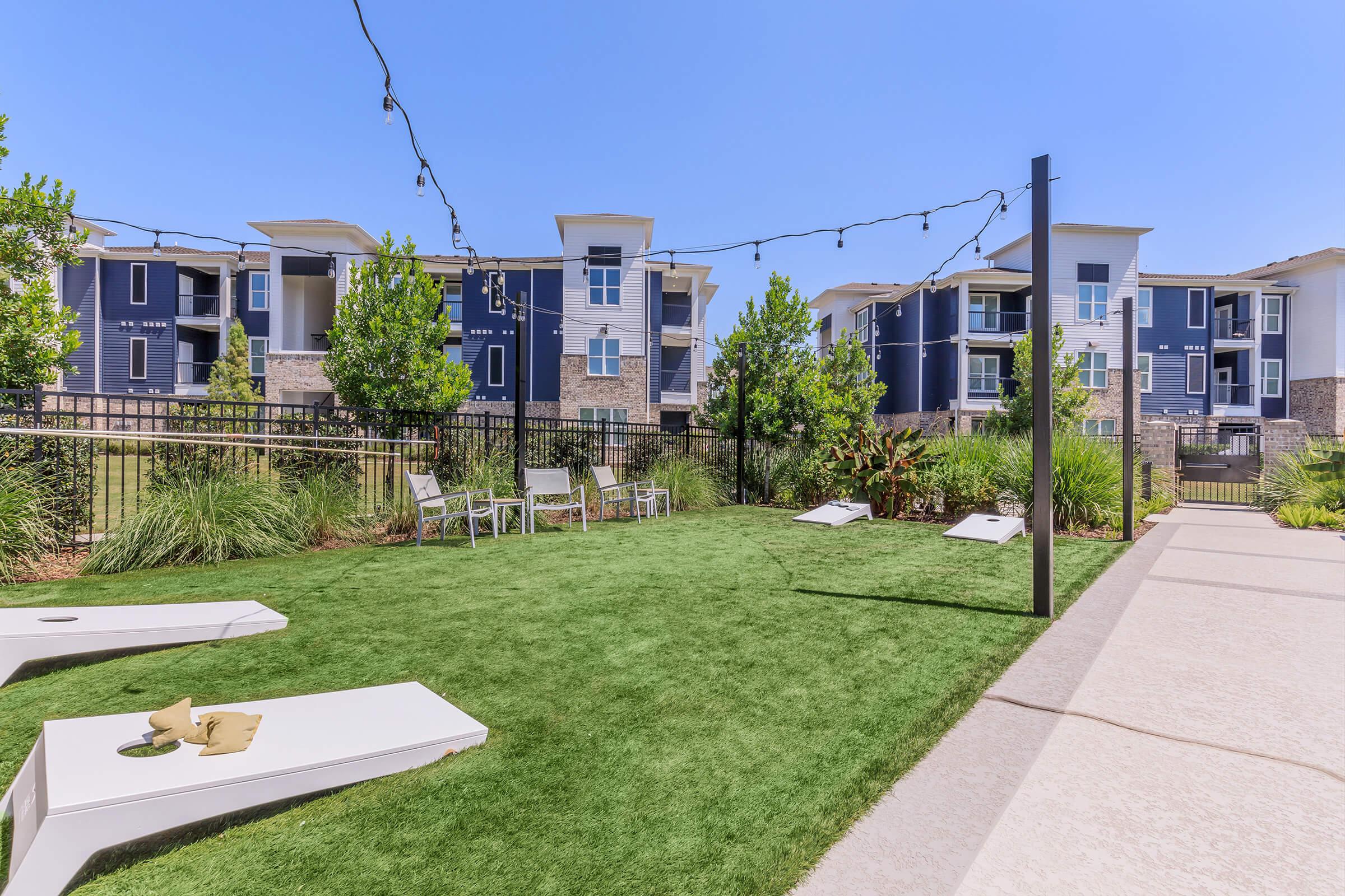 A vibrant outdoor space featuring a well-maintained green lawn with lounge chairs and cornhole games. In the background, modern apartment buildings reflect a clear blue sky, surrounded by shrubs and trees, creating a relaxed atmosphere ideal for leisure and social activities. Decorative string lights enhance the setting.