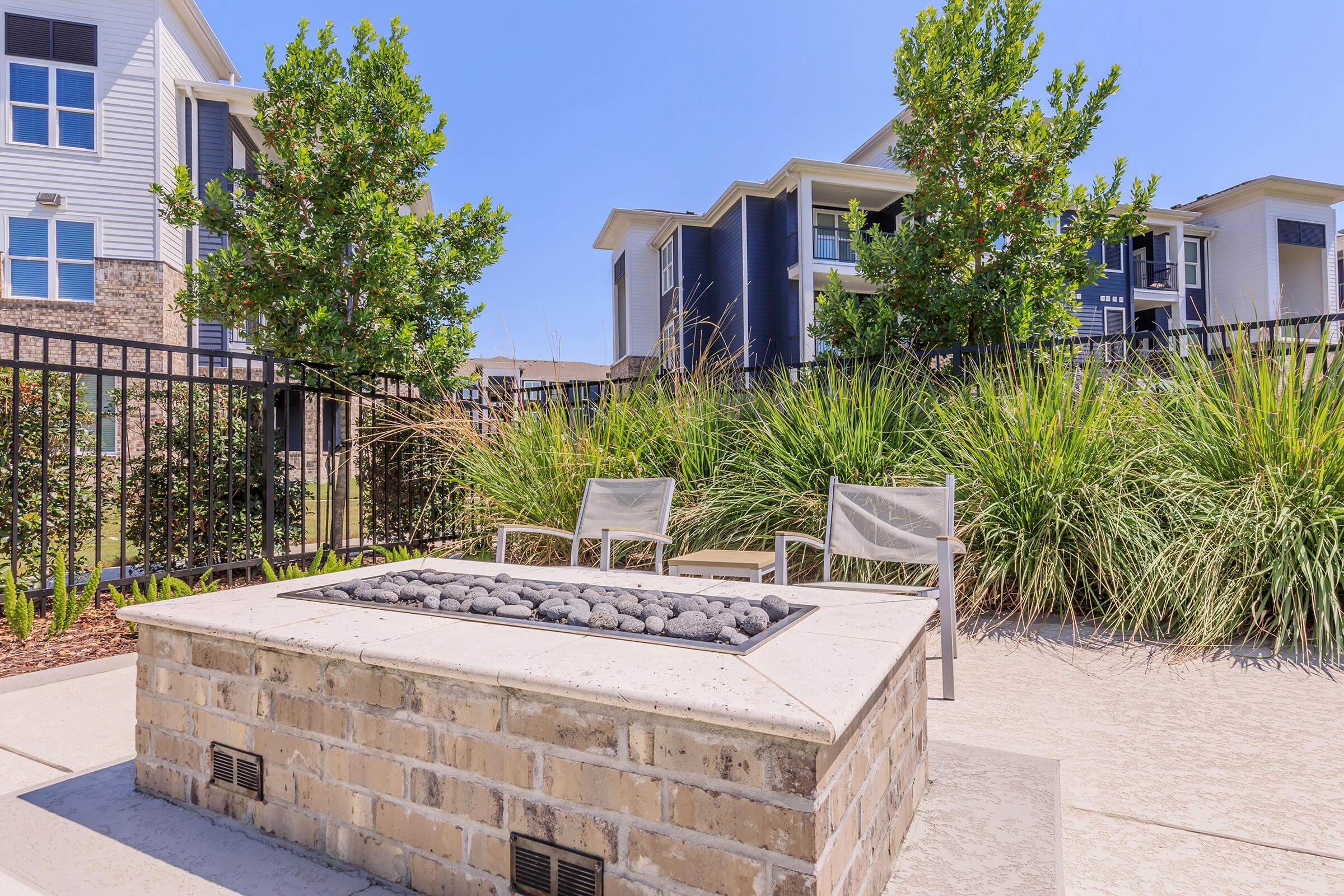 A cozy outdoor space featuring a stone fire pit surrounded by green grass and tall plants. Two chairs are placed nearby, offering a perfect spot for relaxation. In the background, modern apartment buildings are visible under a clear blue sky.