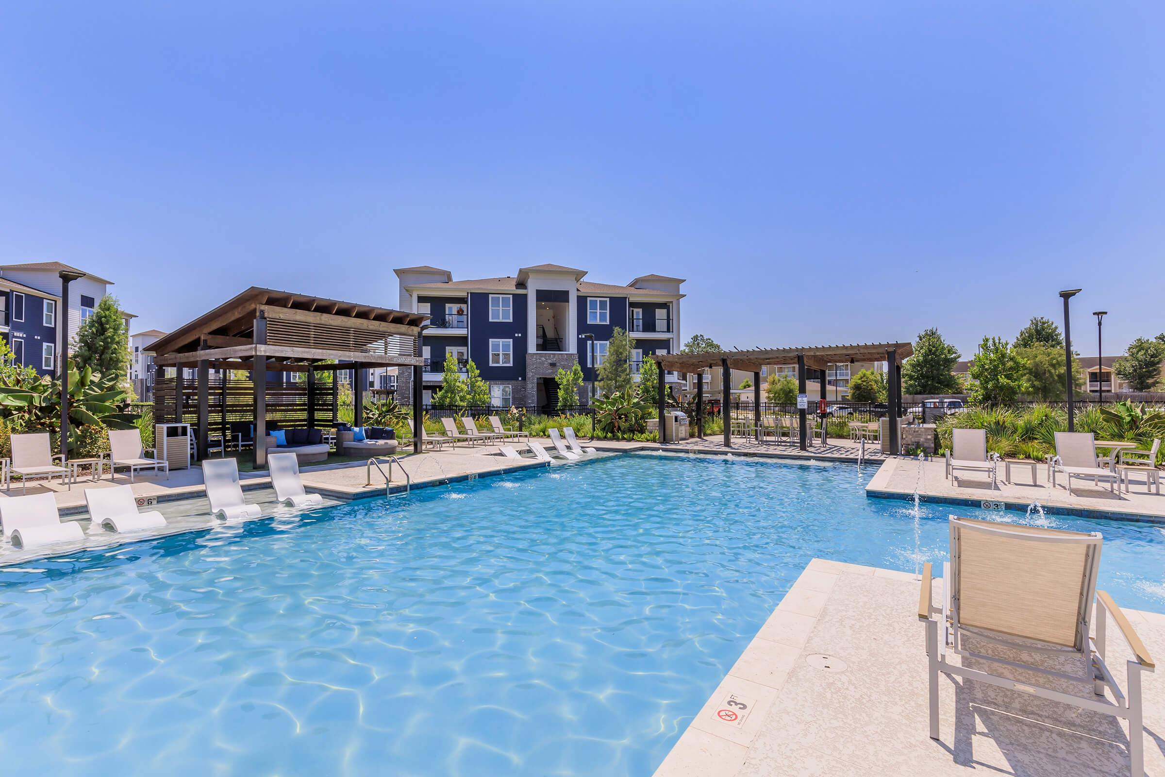 A serene swimming pool surrounded by lounge chairs and shaded cabanas, set against a backdrop of modern apartment buildings. The clear blue water reflects the bright sky, creating a relaxing atmosphere ideal for leisure and relaxation. Lush greenery complements the inviting pool area.
