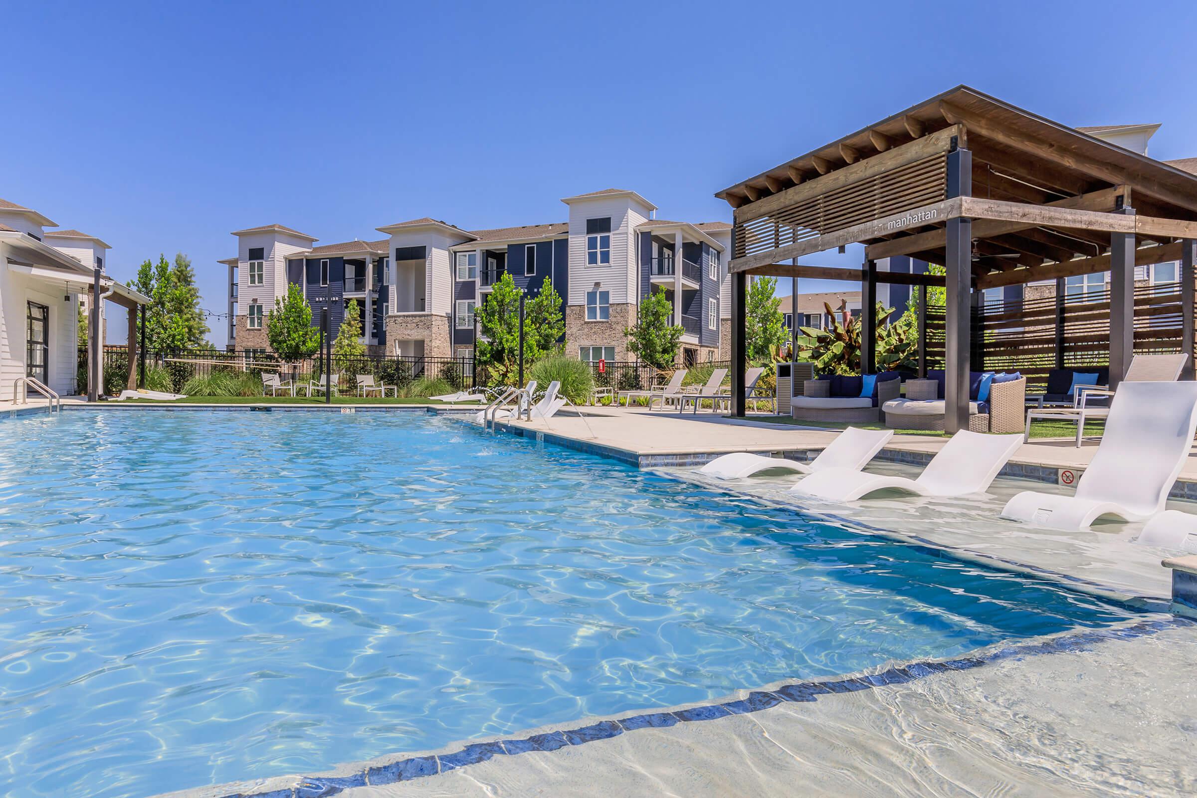 A sunny pool area featuring clear blue water, lounge chairs, and a shaded pergola. In the background, modern apartment buildings are visible surrounded by greenery. The scene conveys a relaxing outdoor atmosphere perfect for leisure and enjoyment.