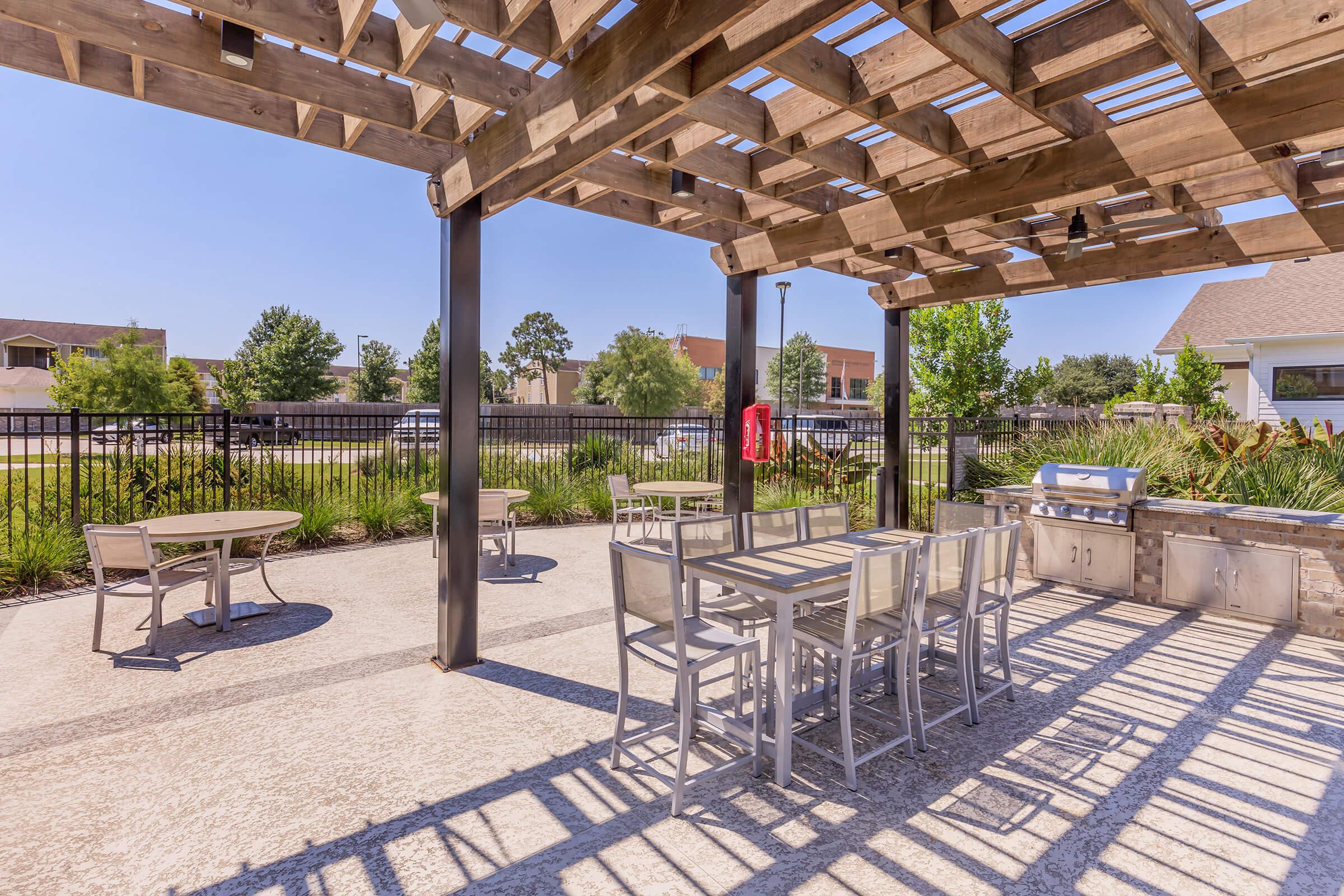 An outdoor patio area featuring a wooden pergola, equipped with metal tables and chairs, surrounded by greenery. A grill area is visible, and the scene is set under a clear blue sky, providing a sunny and inviting atmosphere for gatherings.