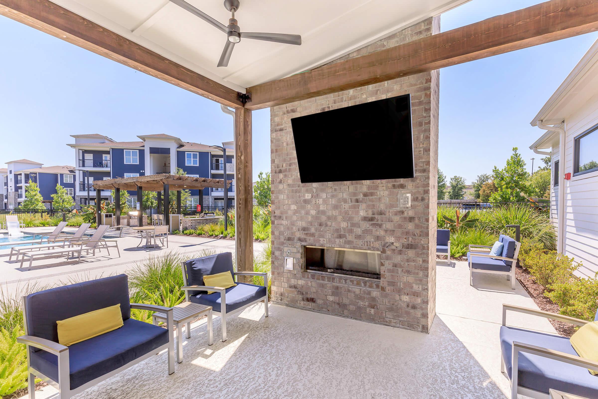 A cozy outdoor lounge area featuring seating with blue cushions and yellow accent pillows, a mounted television on a brick wall, and a fireplace. In the background, there are landscaped grounds with a swimming pool and modern apartment buildings. The scene is bright and sunny.