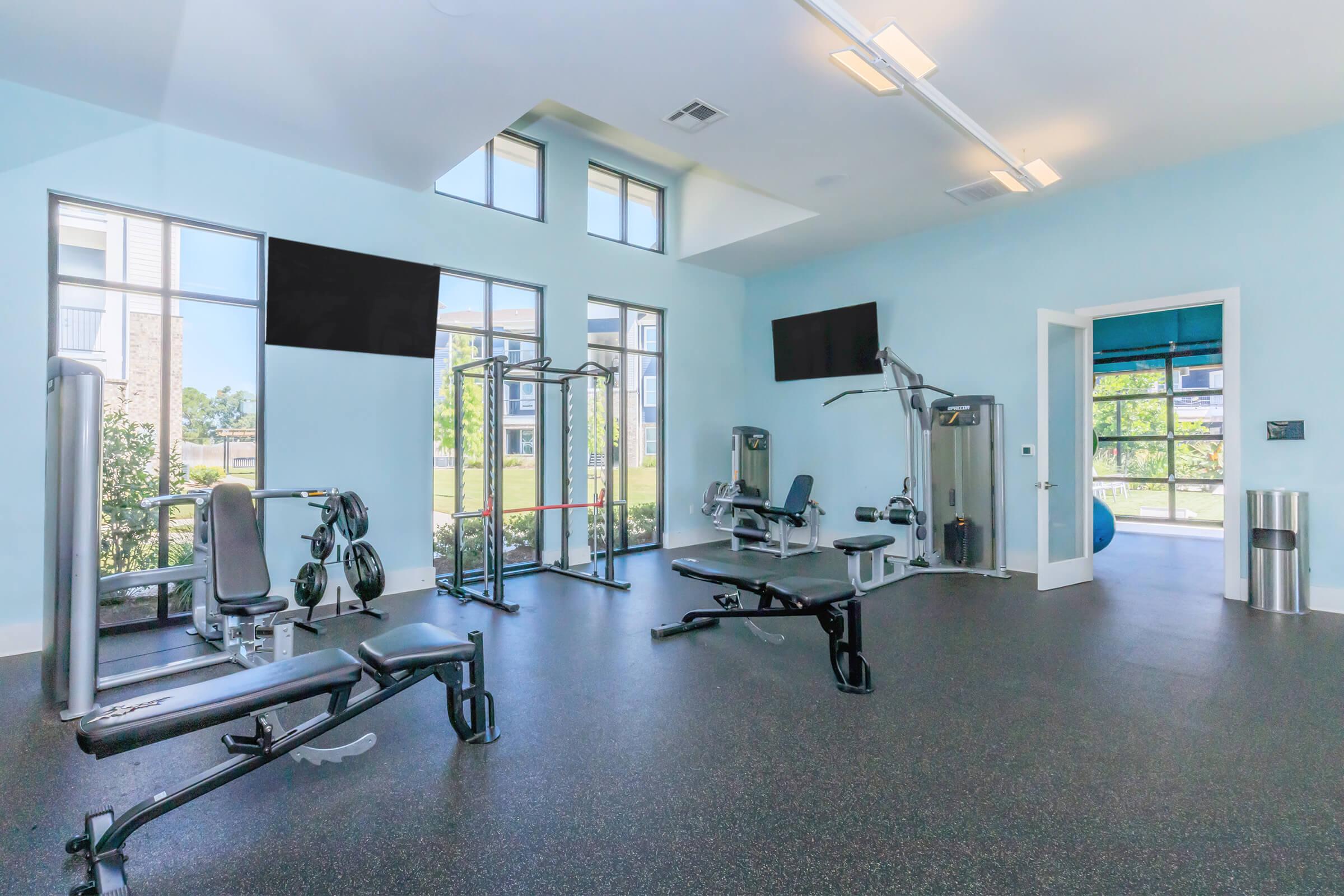 A bright and modern fitness center featuring various exercise machines, including weight machines and free weights. Large windows provide natural light and a view of the outdoors. The flooring is rubberized, and the space is well-organized with a neat, clean appearance.