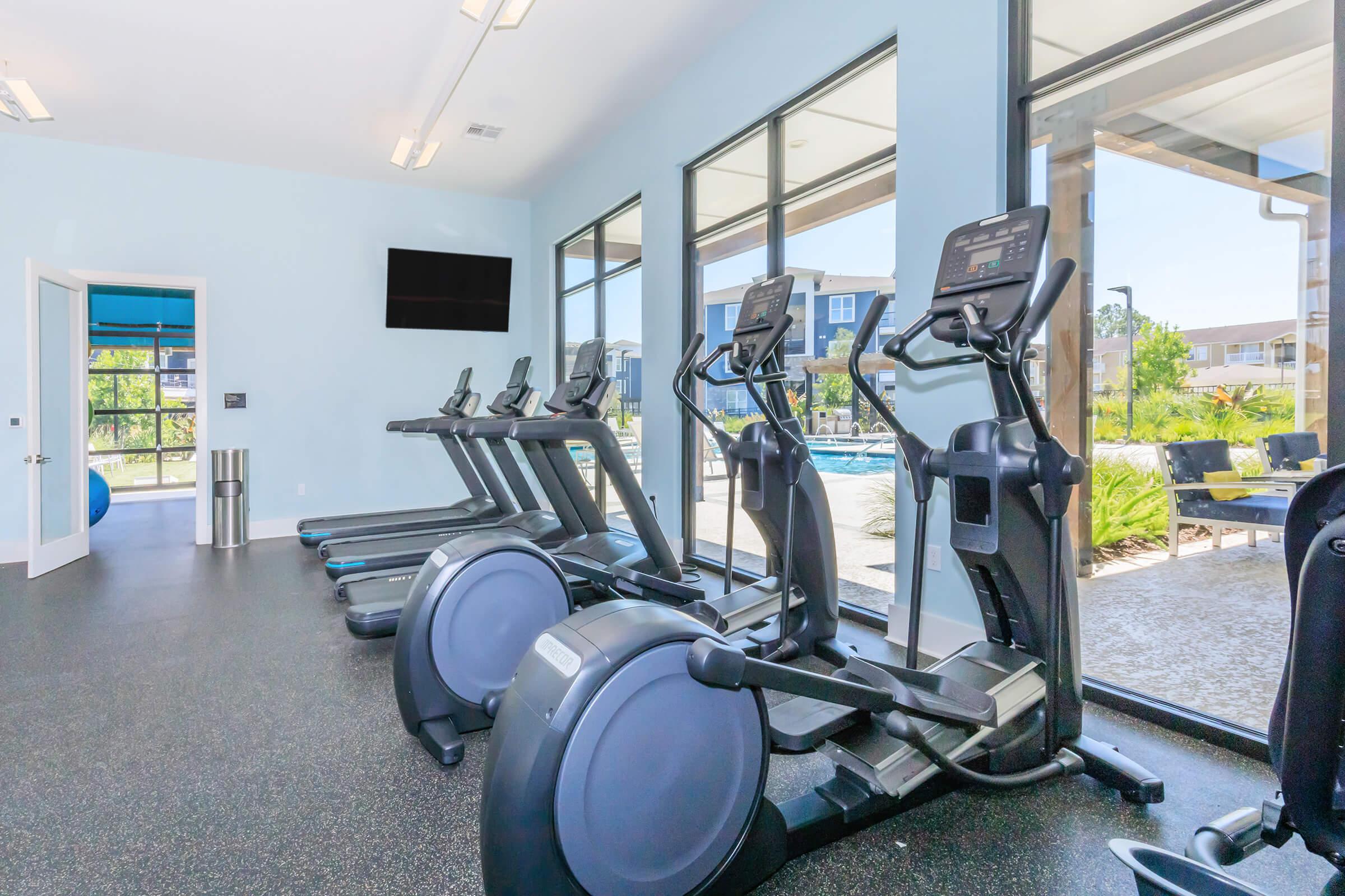 A modern gym interior featuring several treadmills and elliptical machines lined up against large windows that overlook a pool area. The walls are painted light blue, and there’s a flat-screen TV mounted on the wall, creating a bright and inviting workout space.