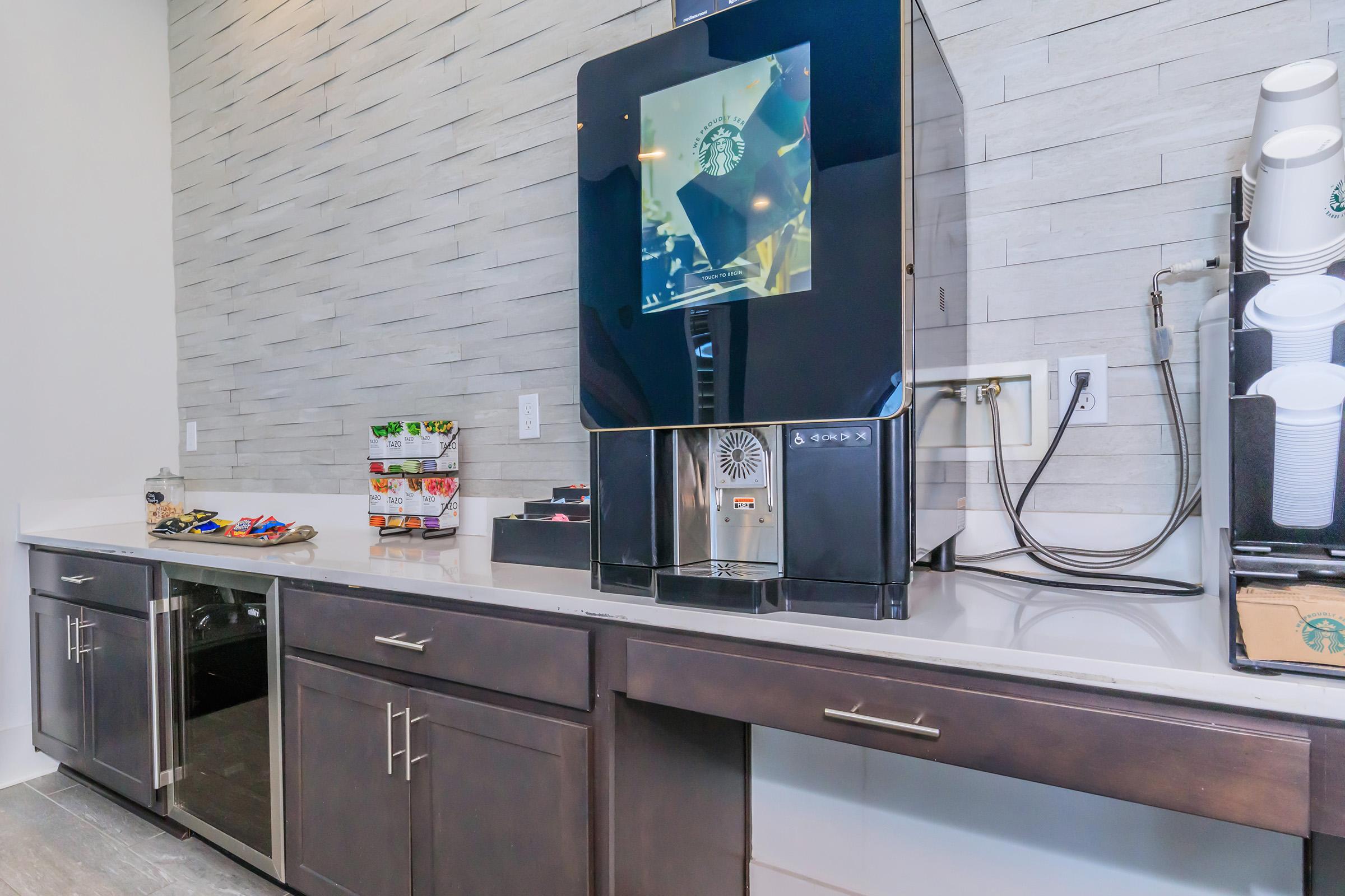 A modern coffee station featuring a black Starbucks coffee machine, snack items in various containers, and a clean countertop with dark cabinetry. The background has textured, light-colored wall tiles, creating a sleek and contemporary look.