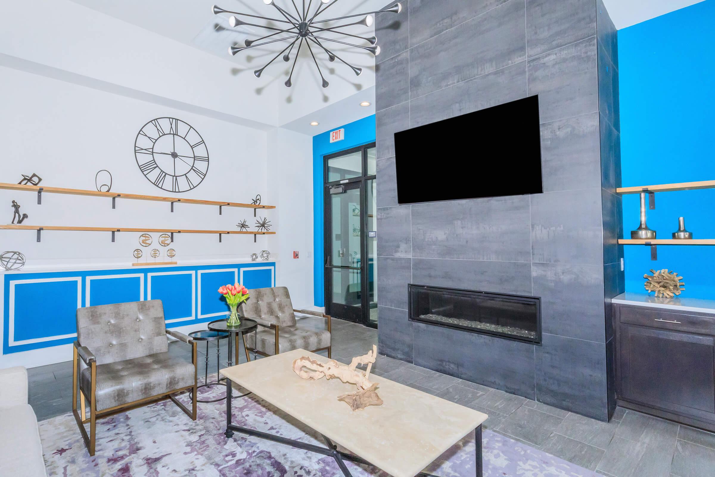 Modern lounge area with a blue accent wall and decorative shelves. A sleek black fireplace is below a large, blank TV screen. Stylish seating includes two gray chairs and a glass-topped coffee table, accompanied by a bouquet of flowers. Large windows provide natural light.