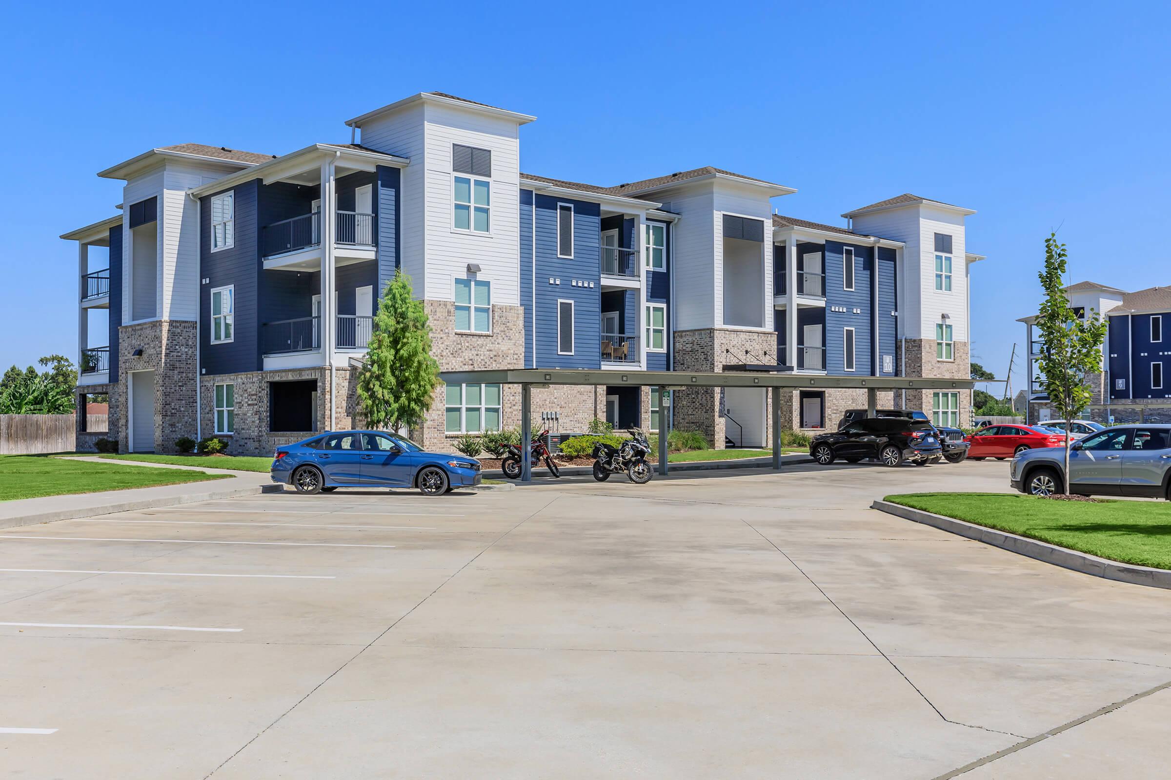 A modern multi-story apartment building with a mix of blue and stone exterior. The building features several balconies and landscaping. In the foreground, there is a paved parking lot with cars and motorcycles parked, along with green grass and trees. Clear blue sky in the background.