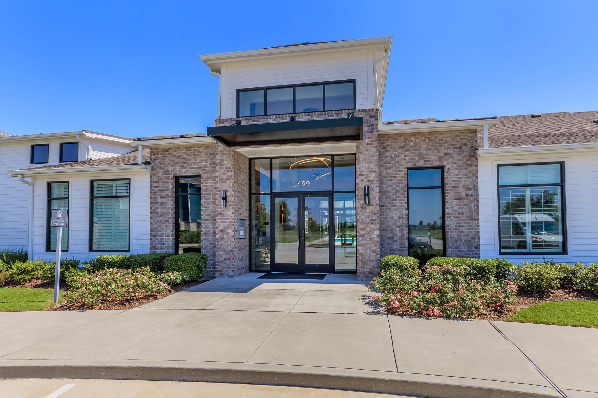 A modern building entrance featuring large glass doors, a flat roof, and a combination of brick and white siding. The facade is flanked by well-maintained landscaping, with a clear blue sky overhead. A sign indicates the address as 1499.