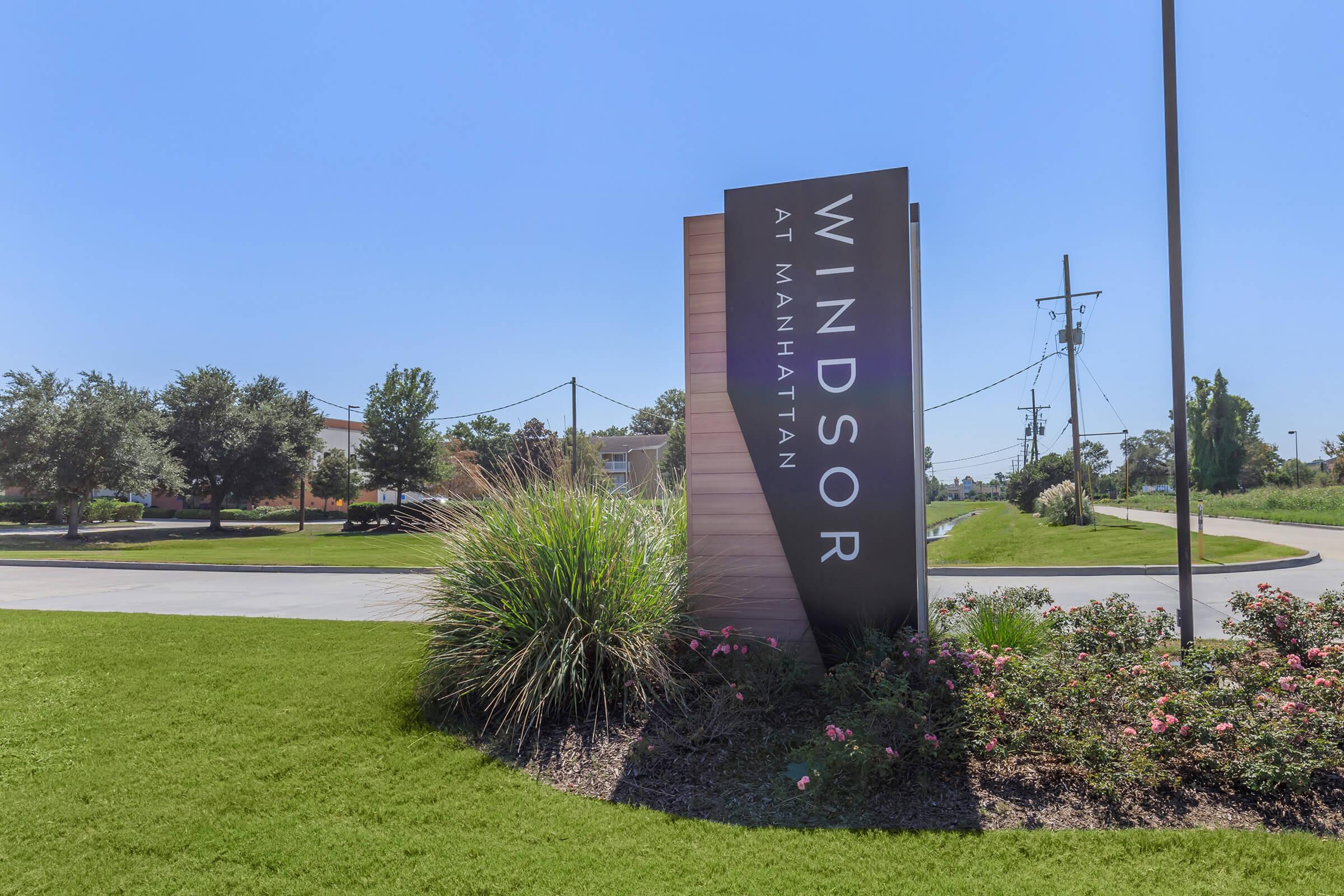 Sign for "Windsor at Manhattan" located near a roadway, surrounded by green grass and colorful landscaping. Clear blue sky in the background.