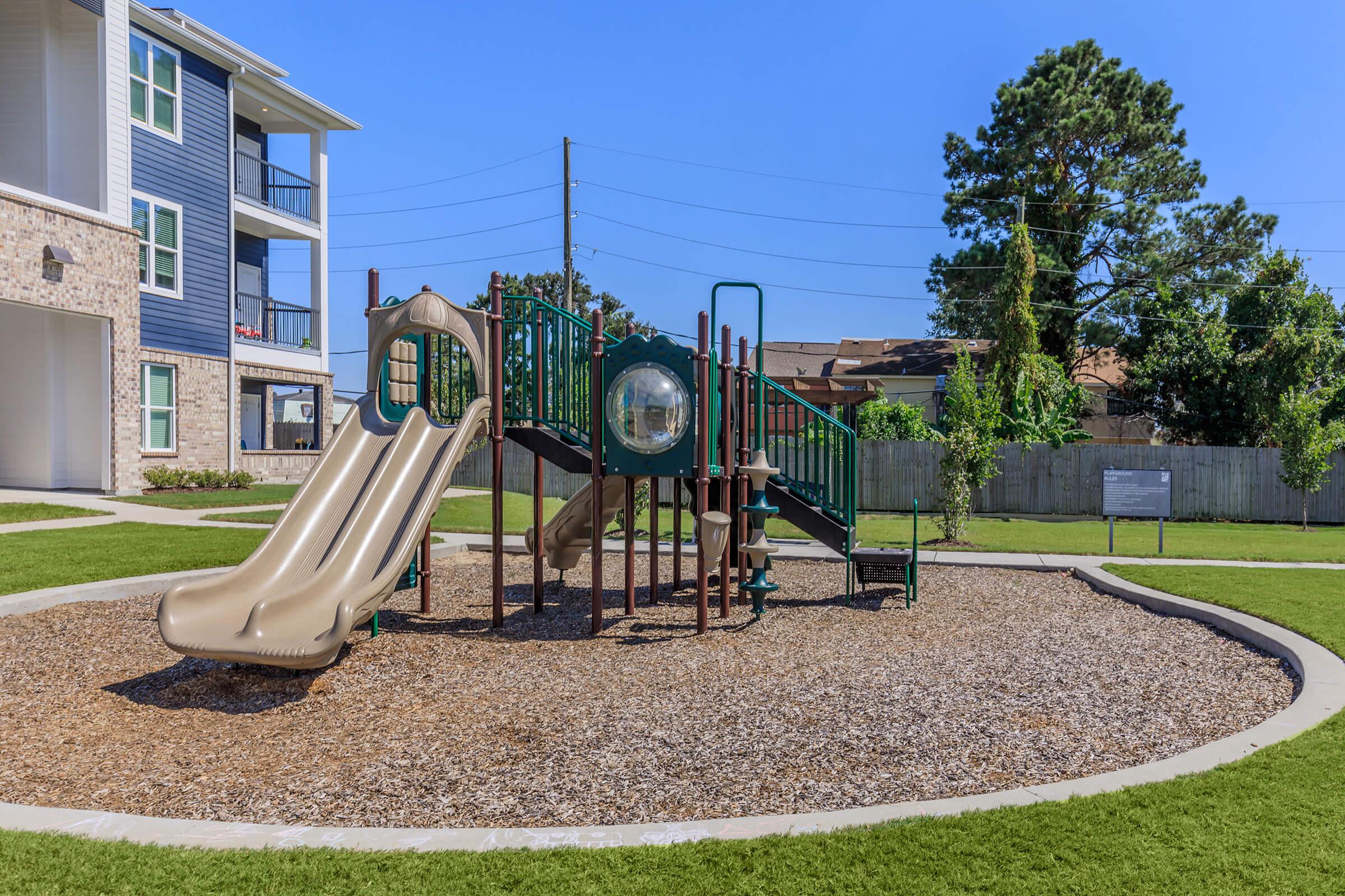 A playground featuring a green climbing structure with a slide and a tunnel, surrounded by a sand-like surface and grass. In the background, there are residential buildings and a wooden fence, under a clear blue sky.