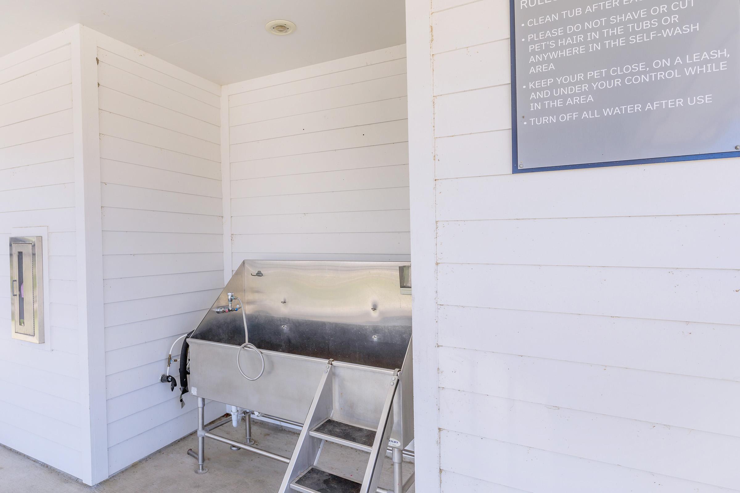 A stainless steel pet wash station with a tub, steps, and signage on the wall providing rules for use. The area features white wooden paneling, creating a clean and organized look for pet owners to groom their animals.