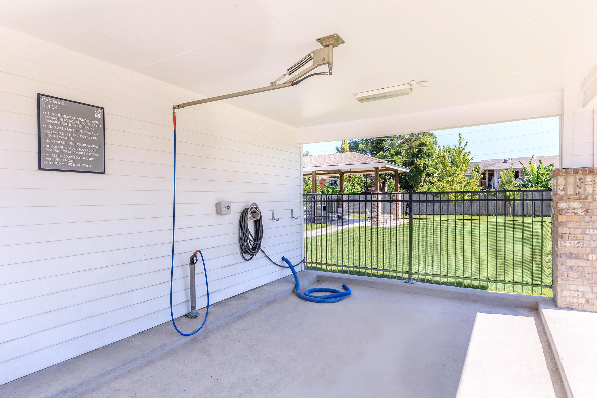 A clean, outdoor pet washing station with a hose mounted on the wall and a blue hose coiled on the ground. Nearby is a grassy area and a fenced yard, with a structure visible in the background, providing a shaded seating area. A sign is mounted on the wall with information or guidelines.