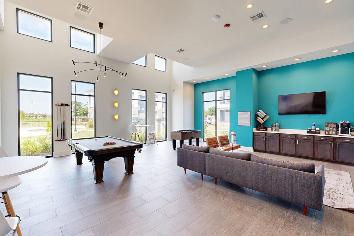 A modern common area with large windows, featuring a billiards table, a foosball table, and a cozy gray sofa. The walls are a mix of white and vibrant teal, with hanging light fixtures and a flat-screen TV. A coffee station is visible, providing a welcoming atmosphere for residents.
