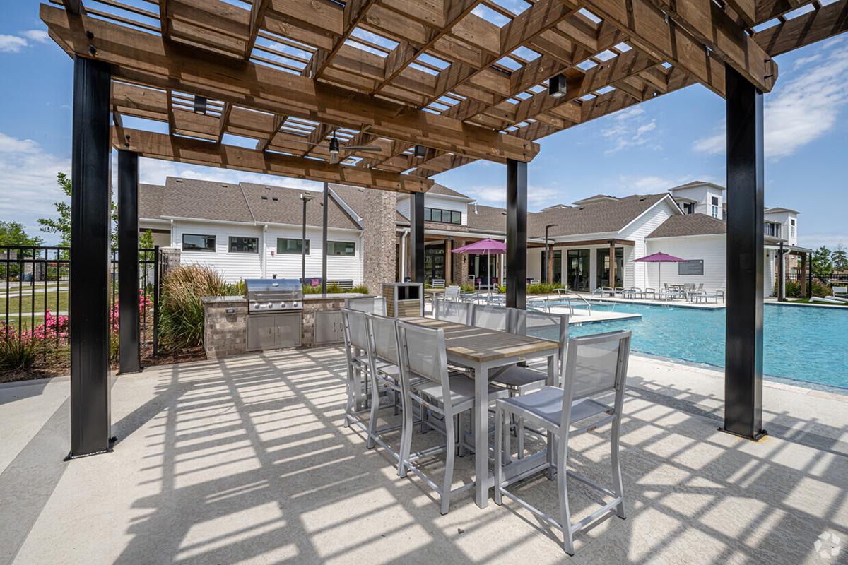 Outdoor kitchen area with a wooden pergola, featuring a grilling station and modern seating. Surrounding a large swimming pool, the space is complemented by vibrant landscaping and lounge chairs, creating a welcoming environment for relaxation and gatherings. Clear blue sky enhances the inviting atmosphere.