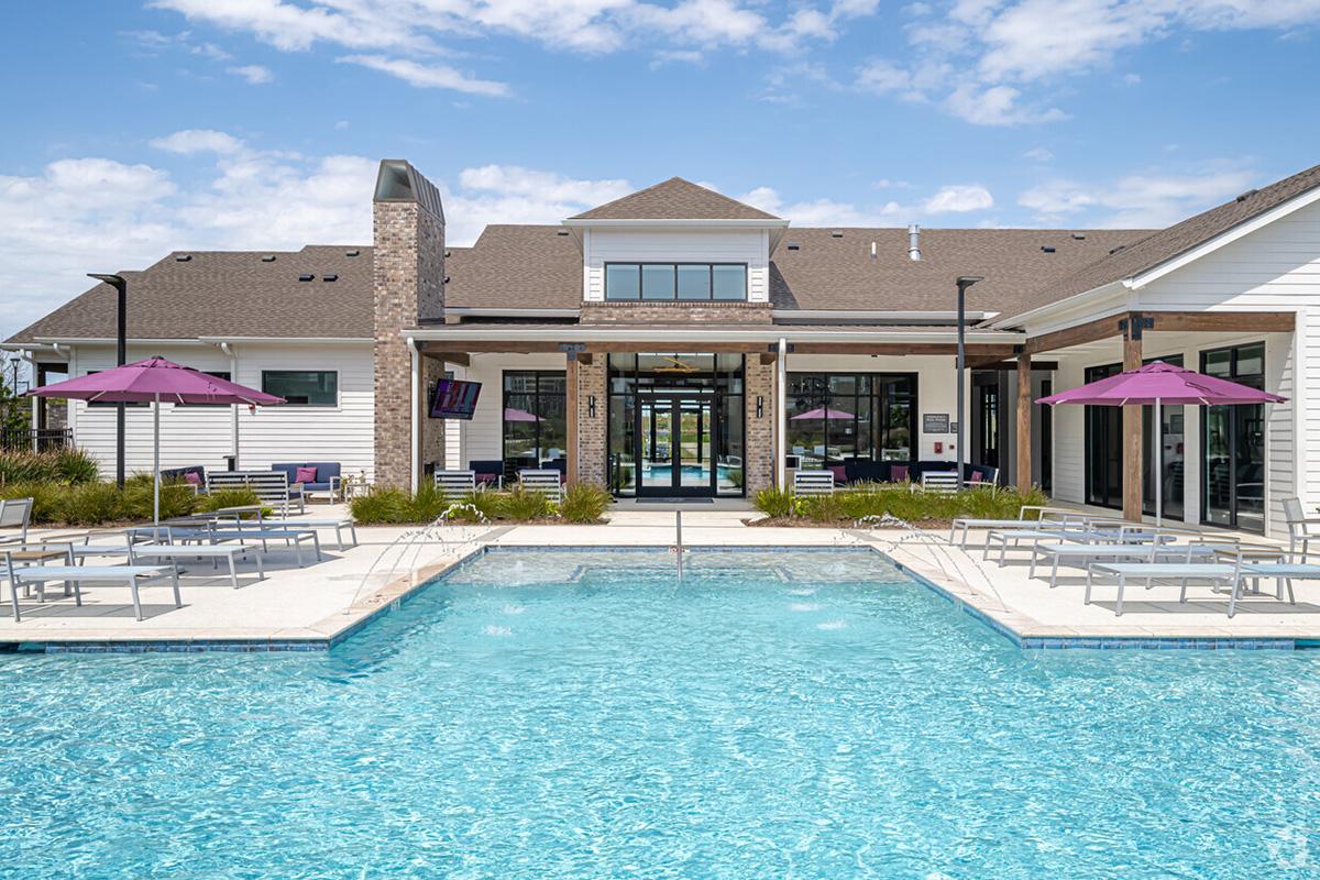 A modern pool area featuring a clear blue swimming pool surrounded by lounge chairs and purple umbrellas. In the background, there is a stylish clubhouse with large windows and a well-maintained landscape, under a bright blue sky with scattered clouds.