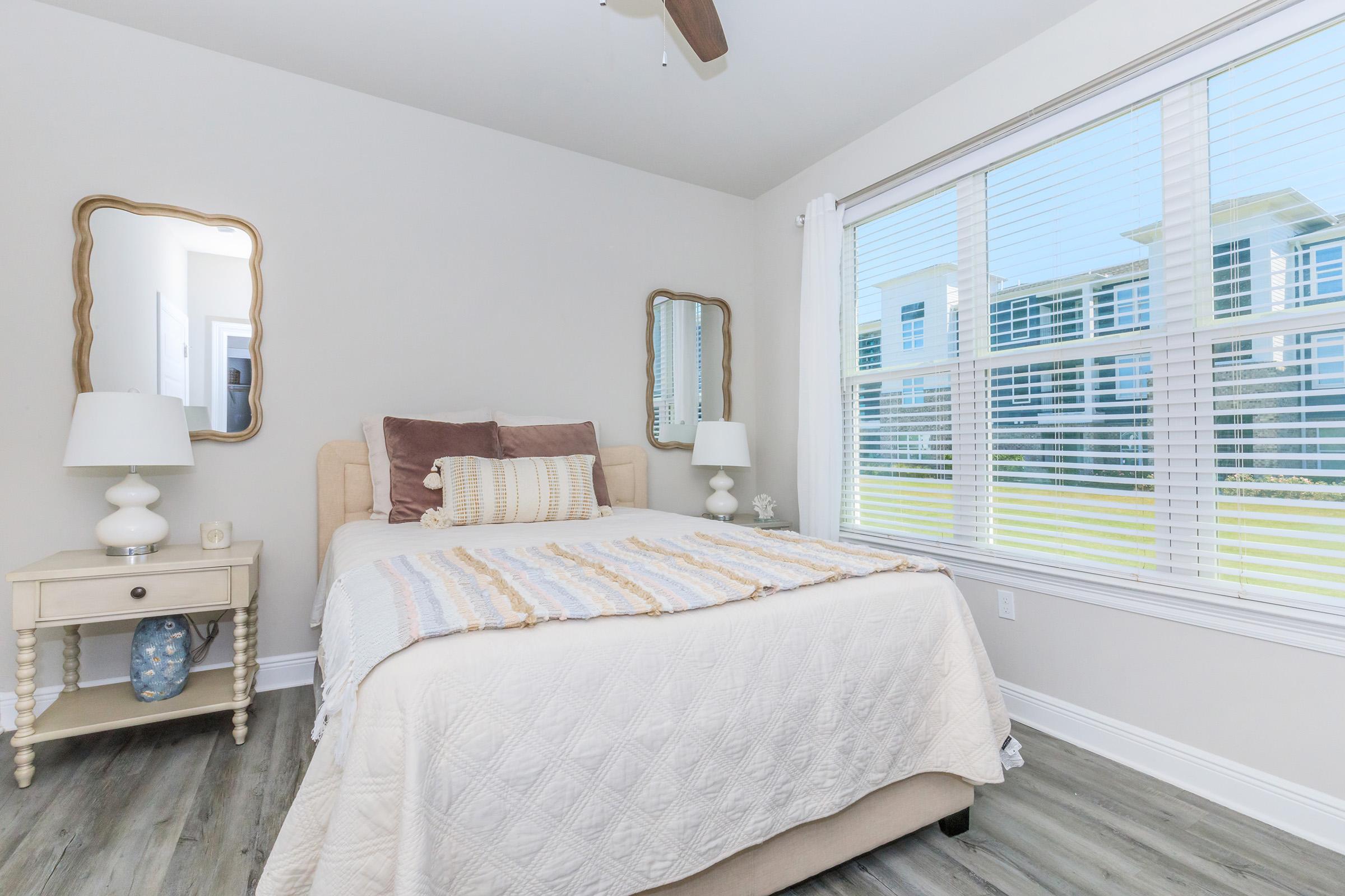 A cozy bedroom featuring a bed with pastel-colored throw pillows and a light quilt. There are two bedside tables with lamps and a decorative mirror above one. Large windows allow natural light to fill the room, offering a view of greenery outside. The flooring is light-colored, enhancing the airy feel.