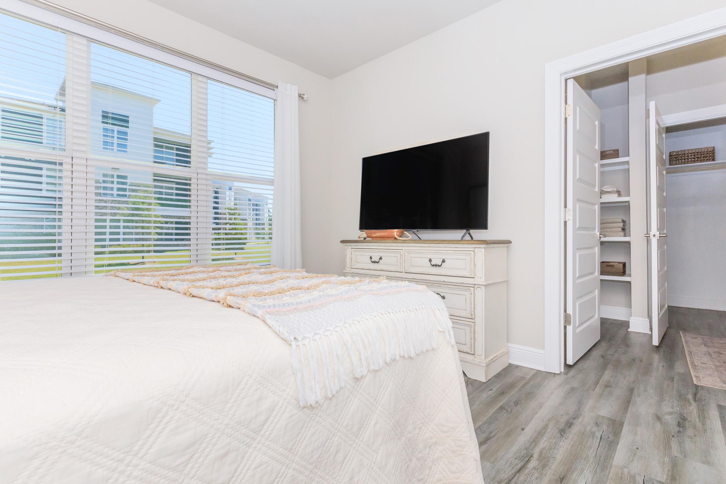 A well-lit bedroom featuring a white quilted bedspread, a large flat-screen TV mounted on a dresser, and a closet with open doors. Soft natural light is coming through the window with blinds, and there's a cozy atmosphere created by the light wood flooring and neutral wall colors.