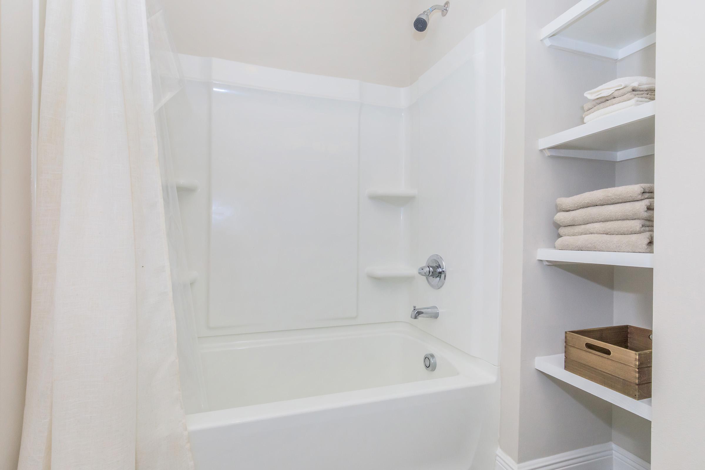 A clean and minimalist bathroom featuring a white bathtub with a showerhead, surrounded by light-colored walls. A soft curtain drapes to one side, and a shelf on the right holds neatly folded towels and a storage basket, creating a serene and organized space.