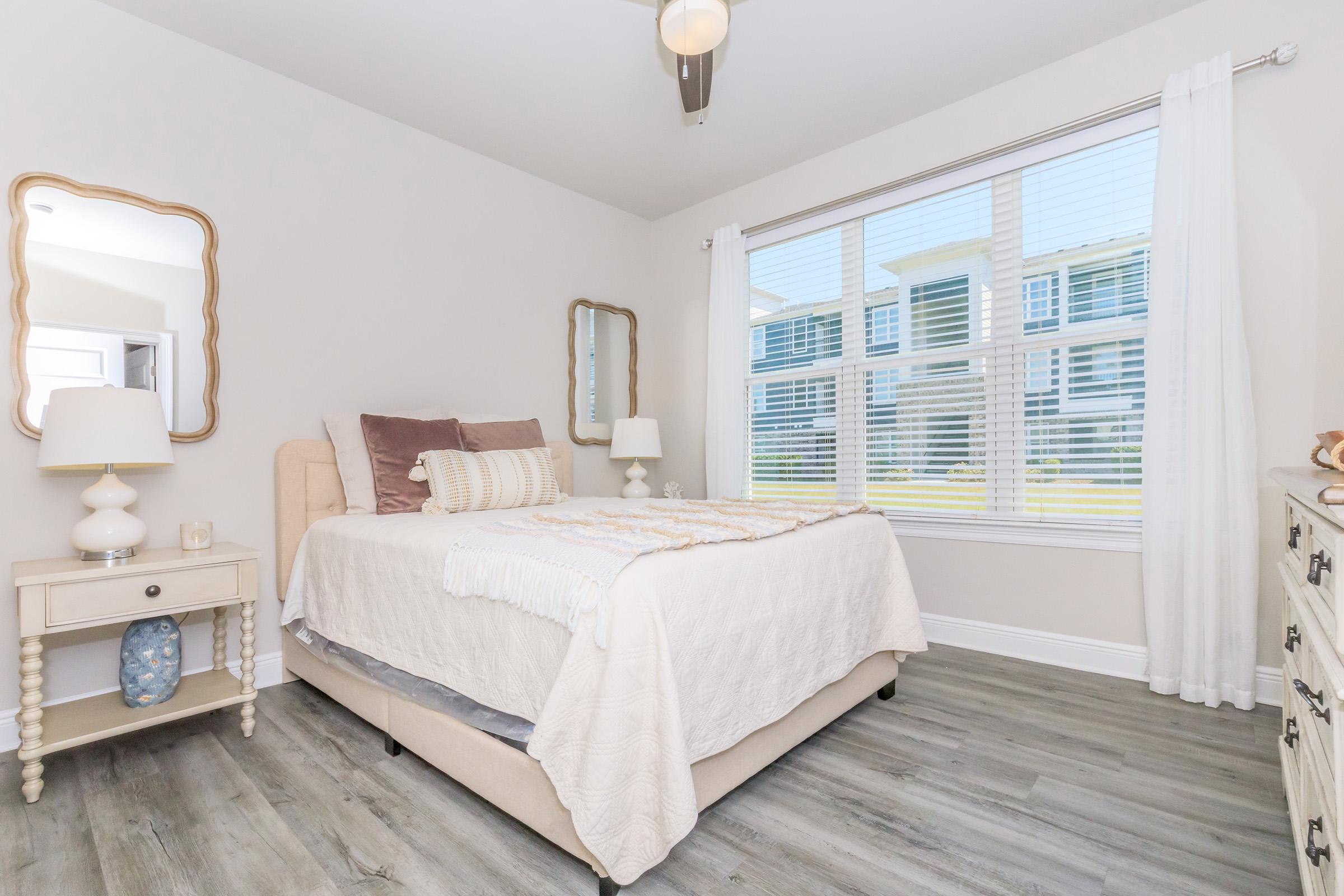 Cozy bedroom featuring a queen-size bed with light bedding, decorative pillows, and a bedside table with a lamp. A large window lets in natural light, complemented by sheer curtains. A stylish mirror and a dresser complete the inviting atmosphere, with soft wood floors adding warmth.