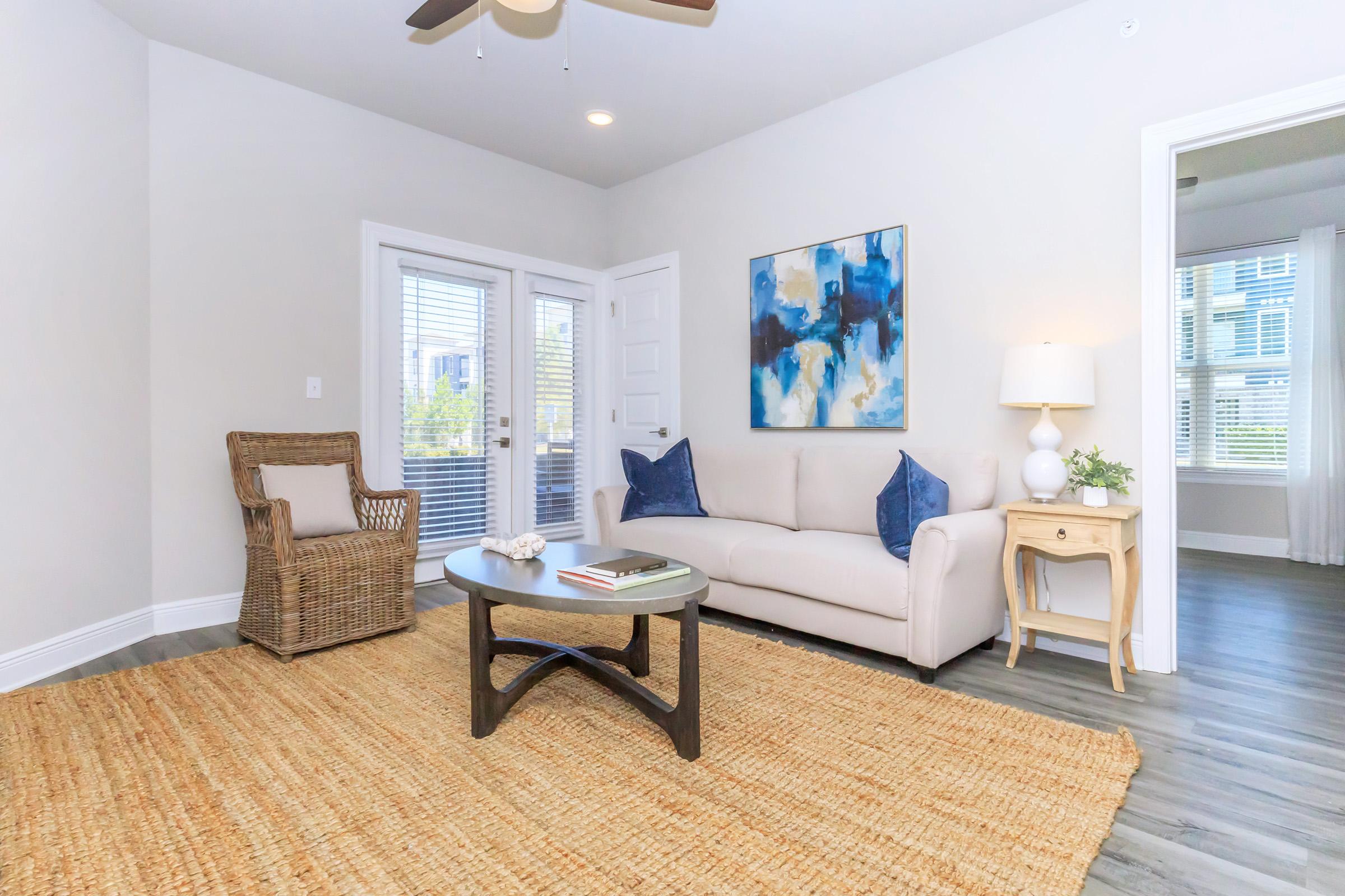 A cozy living room featuring a light gray sofa with blue accent pillows, a round coffee table on a natural fiber rug, and a wicker chair. The walls are painted in a soft color, and a large abstract painting in blue and gold decorates one wall. There are white window shutters letting in natural light.