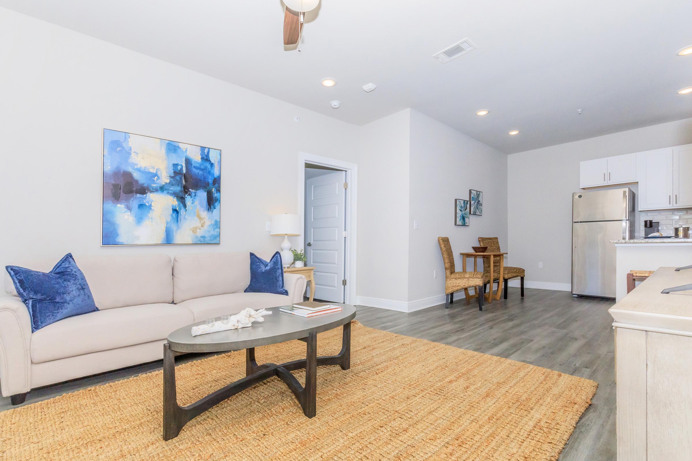 A modern living room featuring a light-colored sofa with blue accent pillows, a circular coffee table, and a woven rug. A dining area with a wooden table and chairs is visible in the background. The space has neutral walls and contemporary lighting, complemented by a colorful abstract painting on the wall.