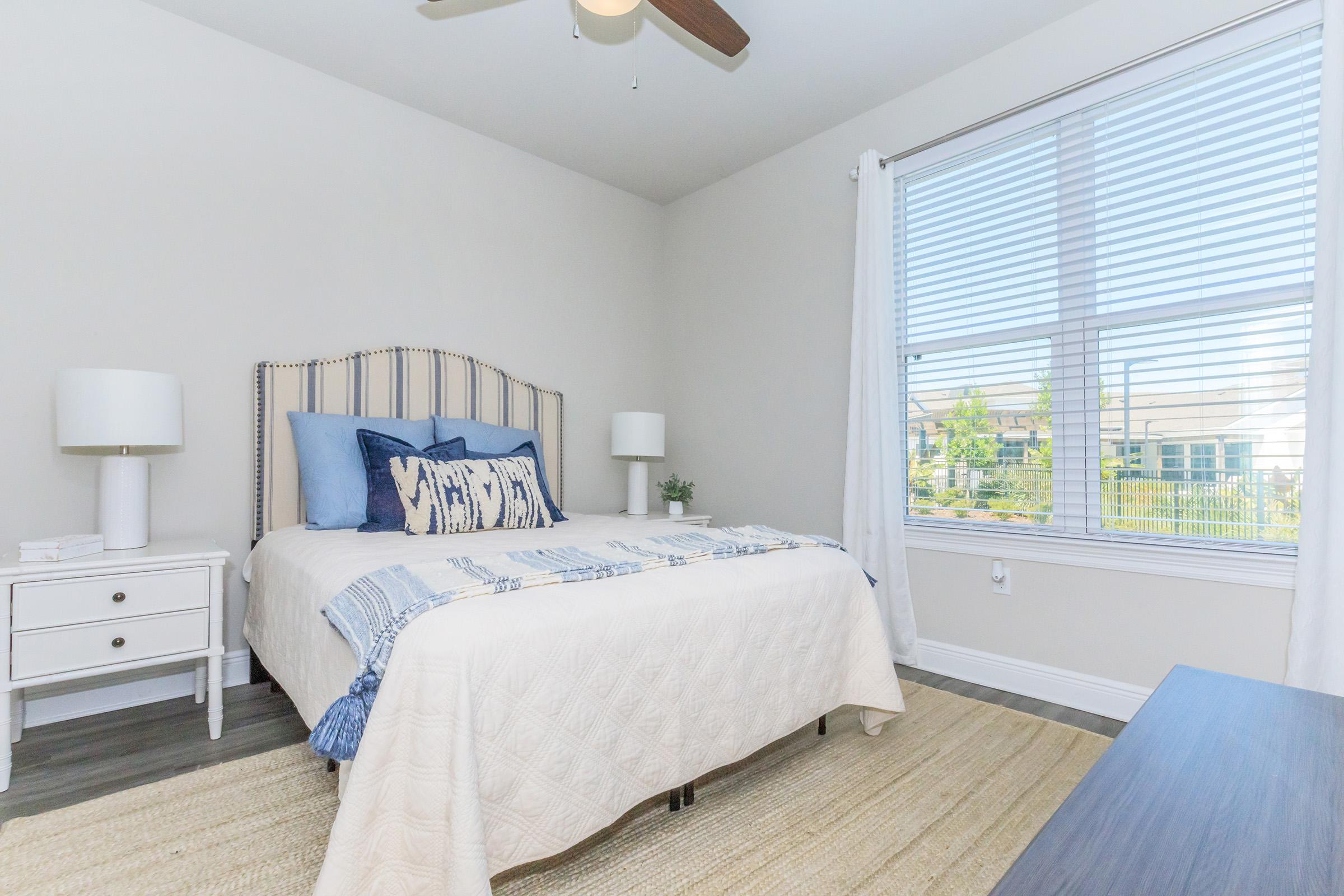 Bright, modern bedroom featuring a neatly made bed with a patterned blue and white throw, two white lamps on bedside tables, and a small indoor plant. Natural light streams in through a window with white blinds, showcasing a view outside. The floor is covered with a light rug, and the walls are painted in a soft color.