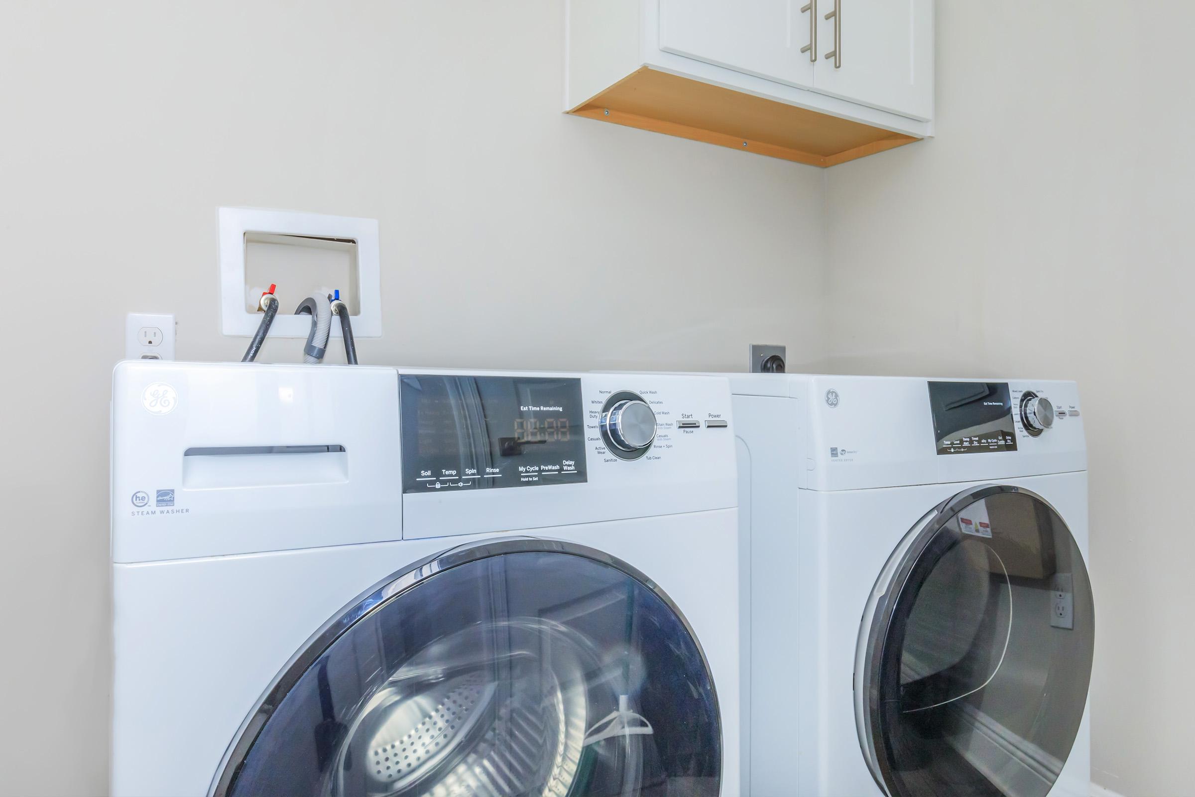 A modern laundry room featuring a pair of white front-loading washing machines side by side, with control panels visible. Above the machines, there is a light-colored cabinet. The wall behind is a neutral shade, providing a clean and organized appearance.