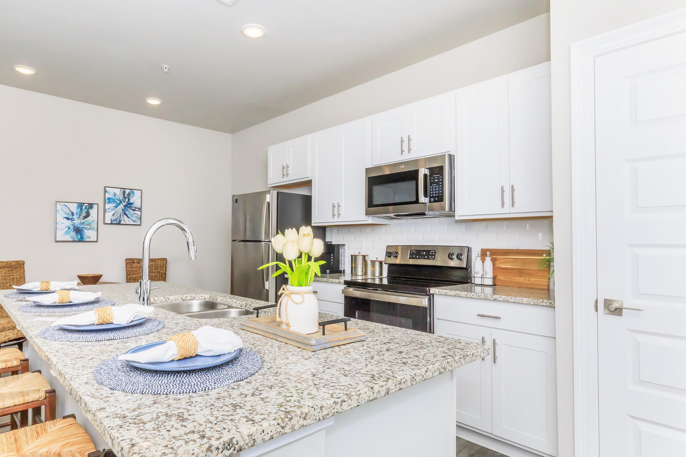 Modern kitchen with granite countertops, white cabinets, stainless steel appliances, and a large sink. The dining area features four place settings with blue and white accents. A vase with white tulips adds a touch of freshness, and there are two framed blue flower artworks on the wall.