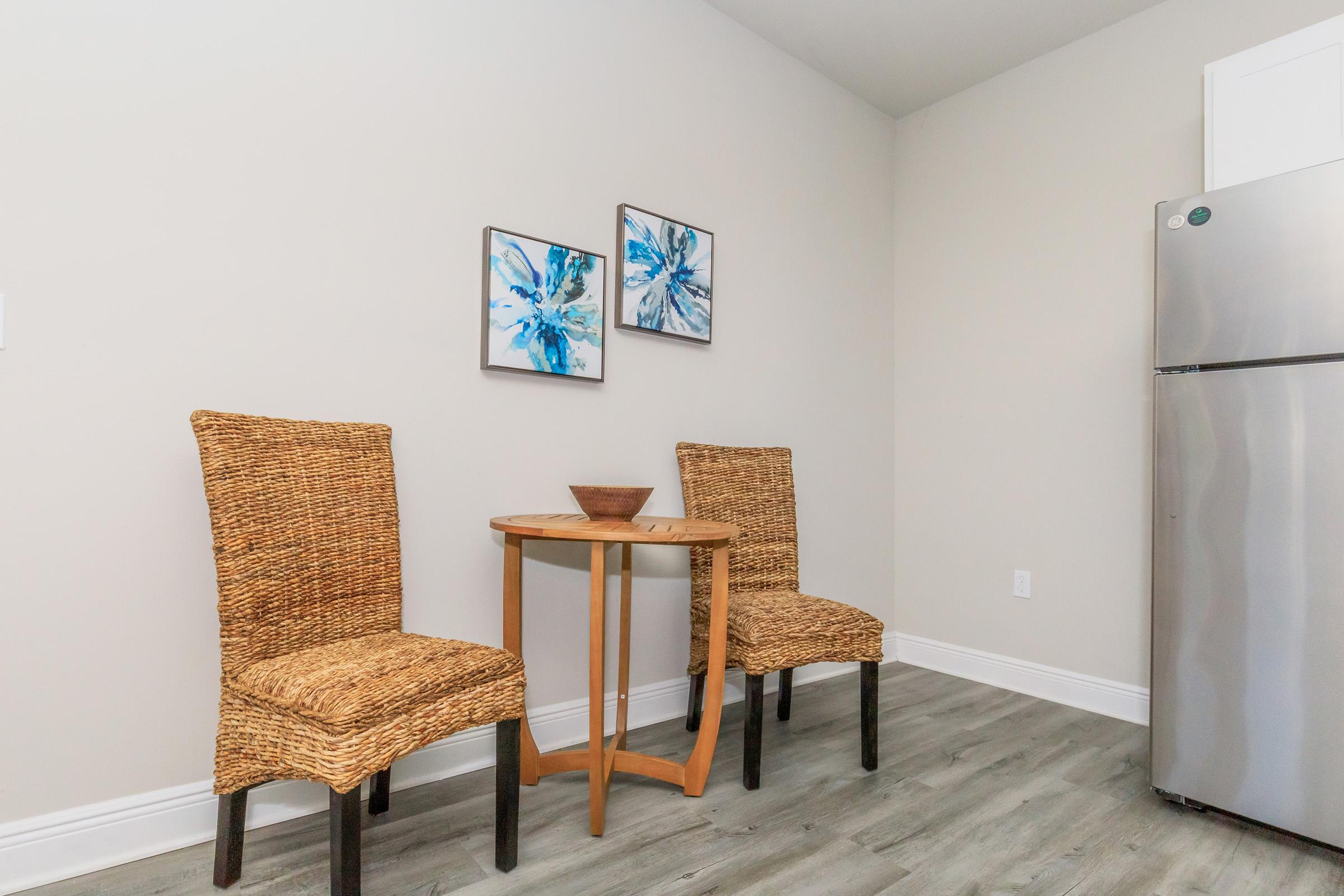 A small dining area featuring two woven chairs and a circular wooden table. On the wall behind, there are two abstract flower paintings in shades of blue. To the right, a stainless steel refrigerator can be seen. The floor has a wood-like finish, and the walls are painted in a light neutral color.