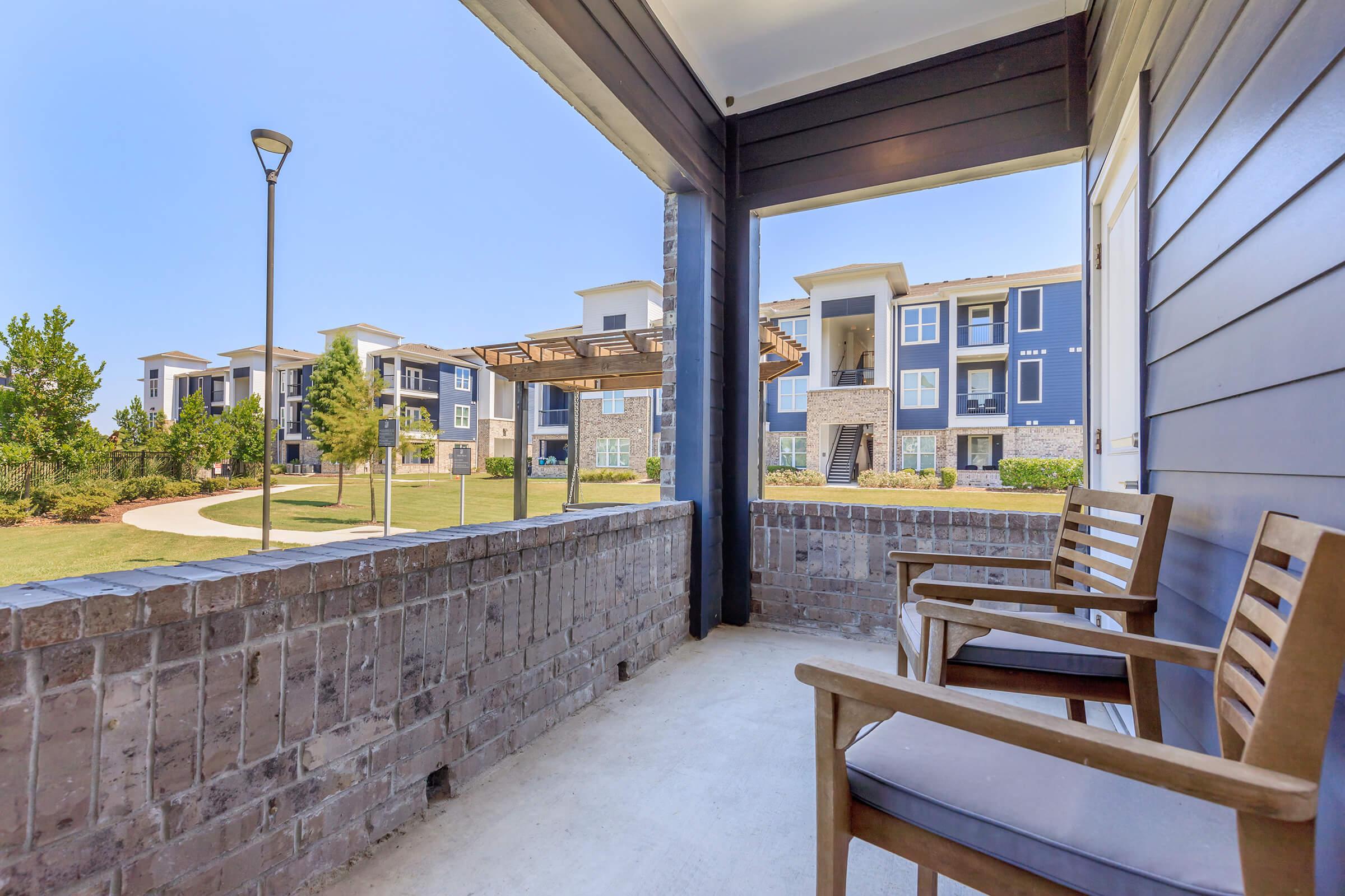 View from a porch overlooking a landscaped area with walking paths and apartment buildings in the background. Two wooden chairs are positioned on the porch, and the scene is set under clear blue skies.