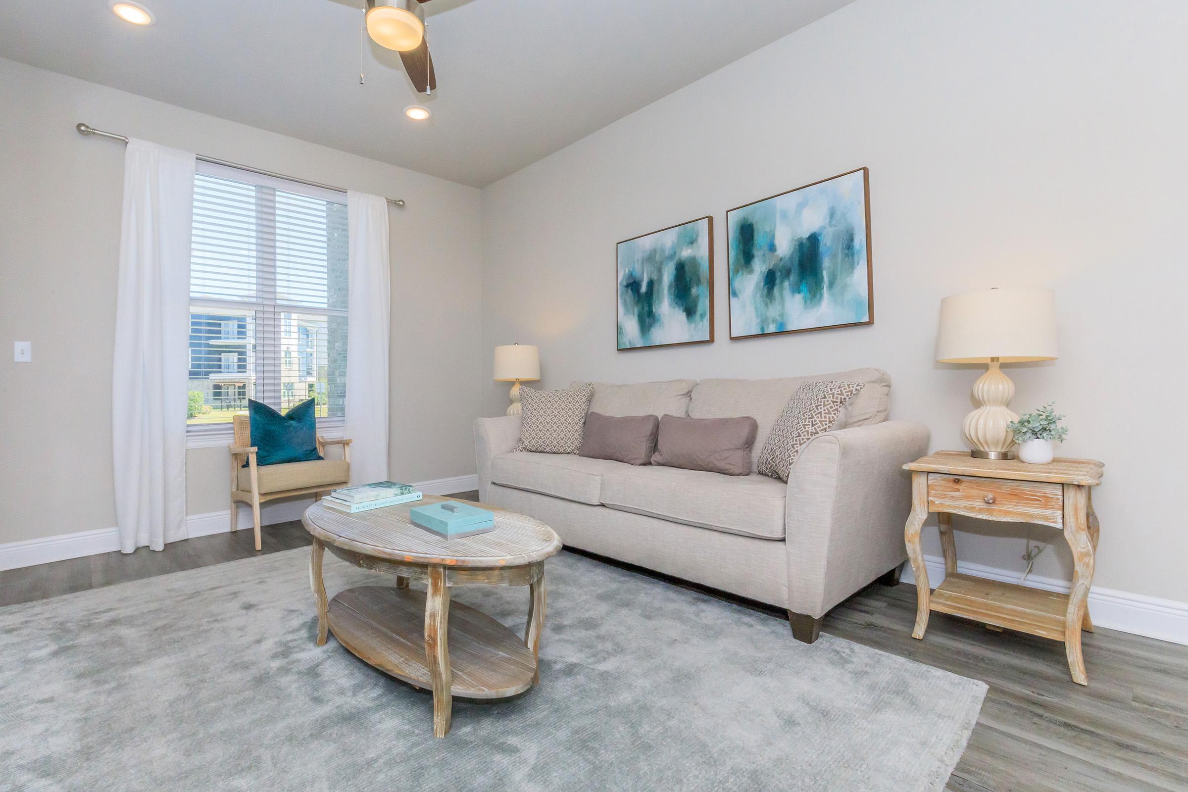 A cozy living room featuring a light beige sofa with decorative pillows, a round wooden coffee table, and a side table with a lamp. Two abstract paintings in blue tones adorn the wall. There is a window with sheer white curtains allowing natural light, and a soft rug covers the wooden floor.