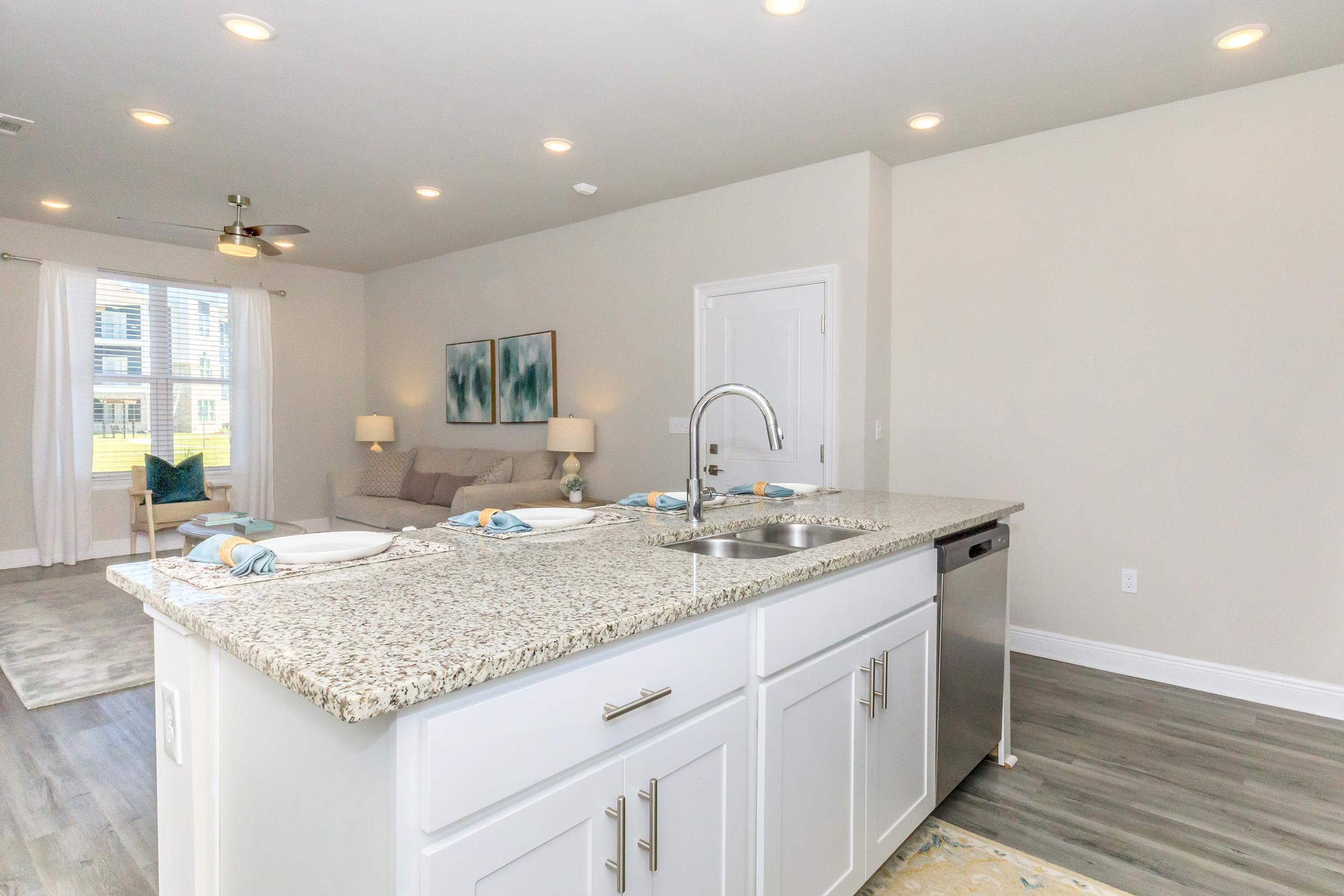 A modern kitchen interior featuring a granite countertop with a sink, white cabinets, and dishes set for dining. In the background, there's a comfortable living area with soft furnishings, large windows, and light-colored walls, creating an inviting ambiance with ample lighting.