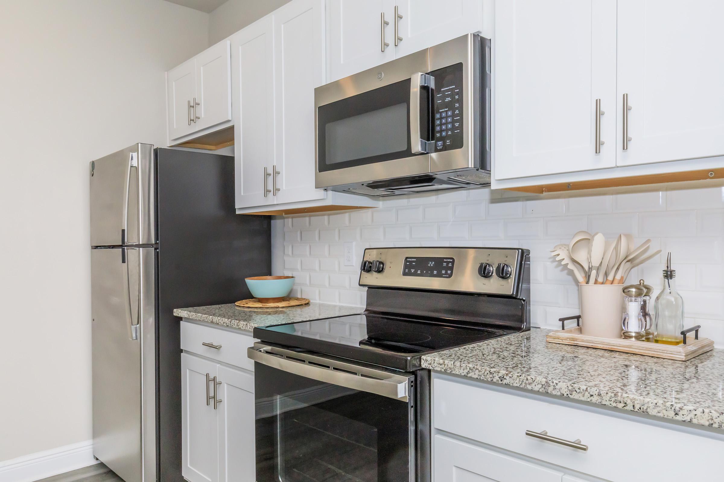 Modern kitchen featuring stainless steel appliances including a refrigerator, microwave, and oven. The countertops are made of granite, and there is a bowl on the counter along with cooking utensils. White cabinets provide a clean, sleek look, complemented by a backsplash of white tiles.