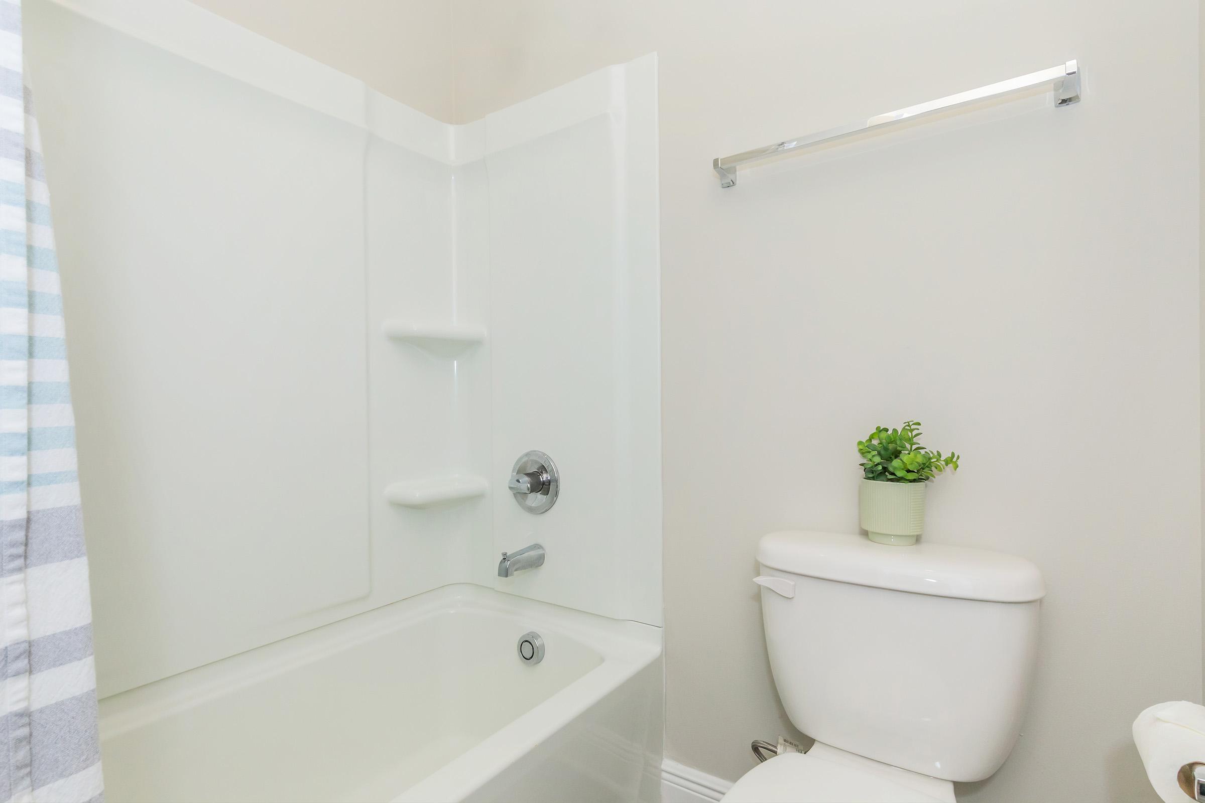 A clean bathroom featuring a shower-tub combination, a toilet, and a small potted plant on the toilet tank. Neutral-colored walls and a striped shower curtain add to the decor. A towel bar is mounted on the wall.