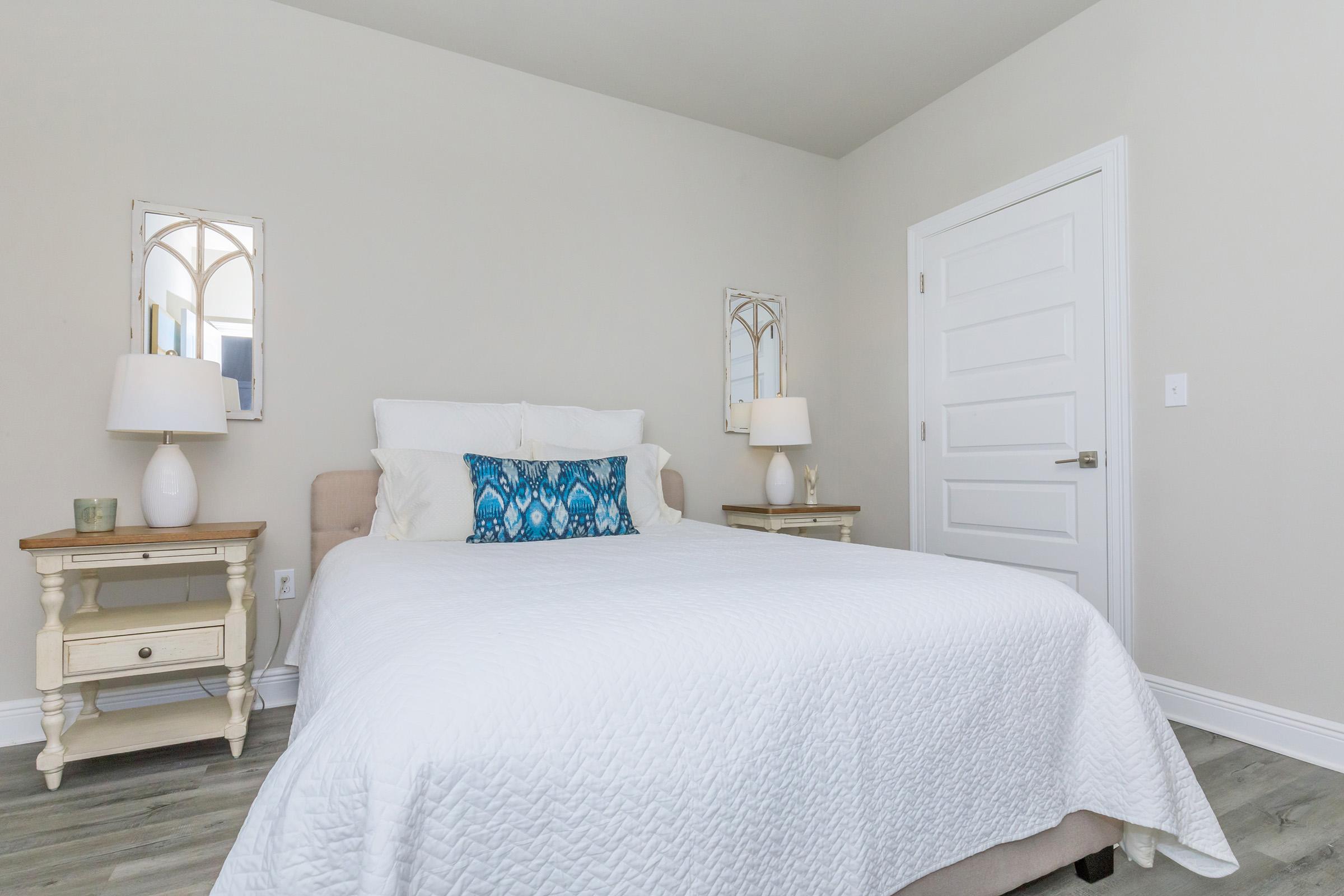 A neatly arranged bedroom featuring a light-colored wall, a double bed with a white quilt, and decorative pillows. Two bedside tables with lamps are positioned on either side of the bed. Mirrors are mounted above the tables, and a white door is visible in the background.
