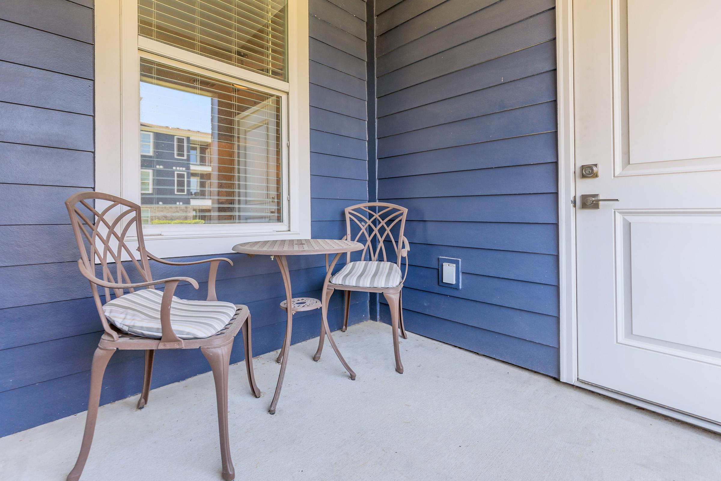 A cozy porch area featuring two metal chairs with cushions and a small round table, positioned near a door. The wall has horizontal blue siding, and a window lets in natural light.