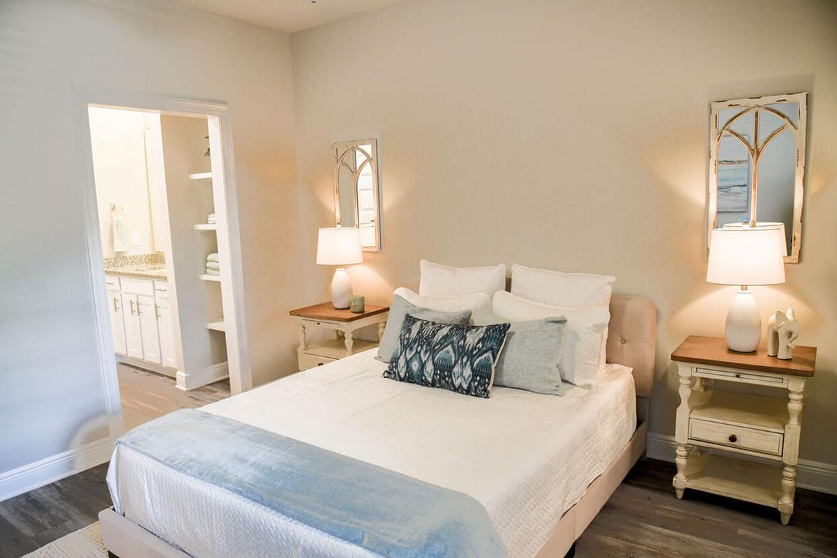A cozy bedroom featuring a neatly made bed with white linens and decorative pillows, two bedside tables with lamps, and wall mirrors. There is a softly lit entrance to a bathroom visible in the background, and light-colored walls create a warm and inviting atmosphere.