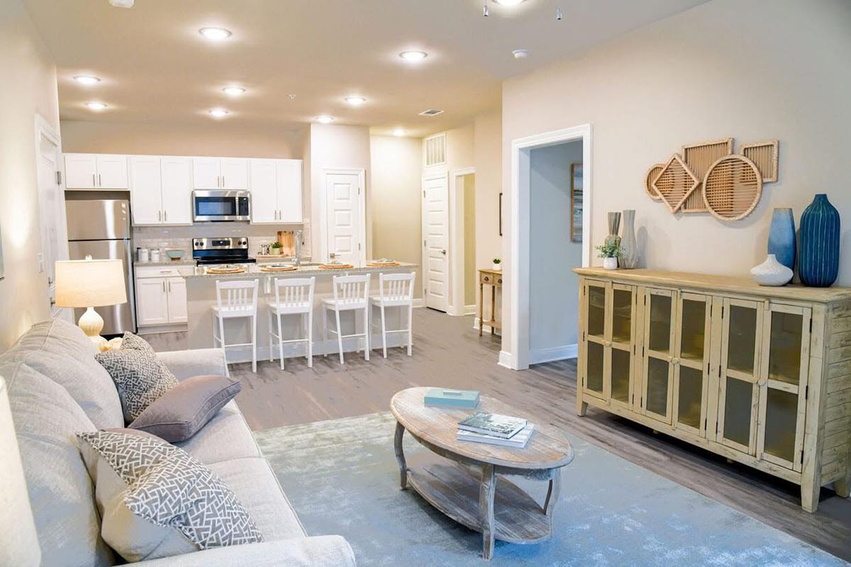 A modern, open-concept living space featuring a cozy living room with a light-colored sofa and decorative pillows, a coffee table, and a stylish cabinet. The kitchen area includes white cabinetry and stainless-steel appliances. Soft lighting and neutral colors create a warm, inviting atmosphere.