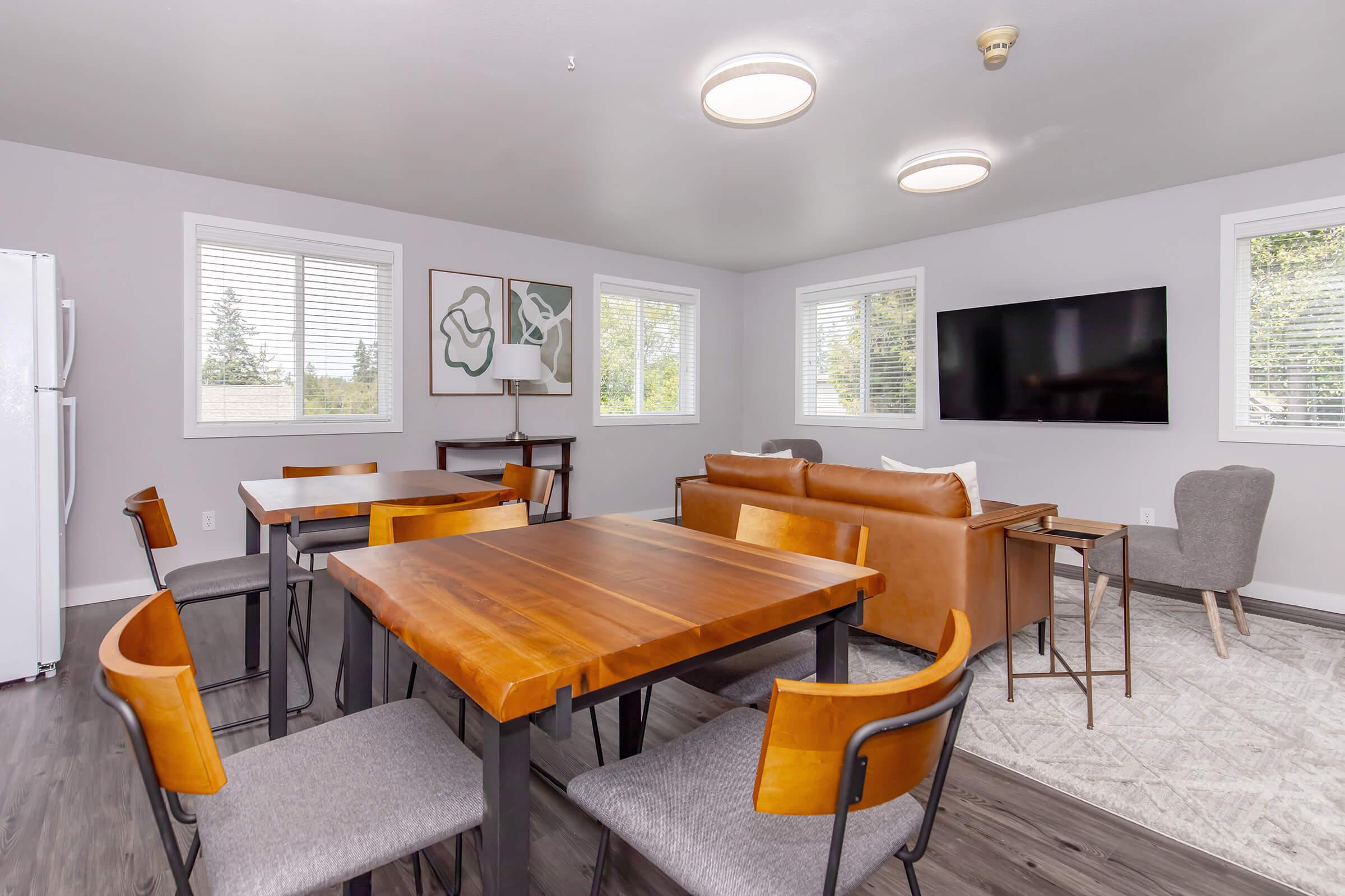 A modern living space featuring a dining area with wooden tables and chairs, a cozy brown leather sofa, and a gray armchair. The room has large windows letting in natural light, a TV mounted on the wall, and minimalistic decor, creating a stylish and inviting atmosphere.