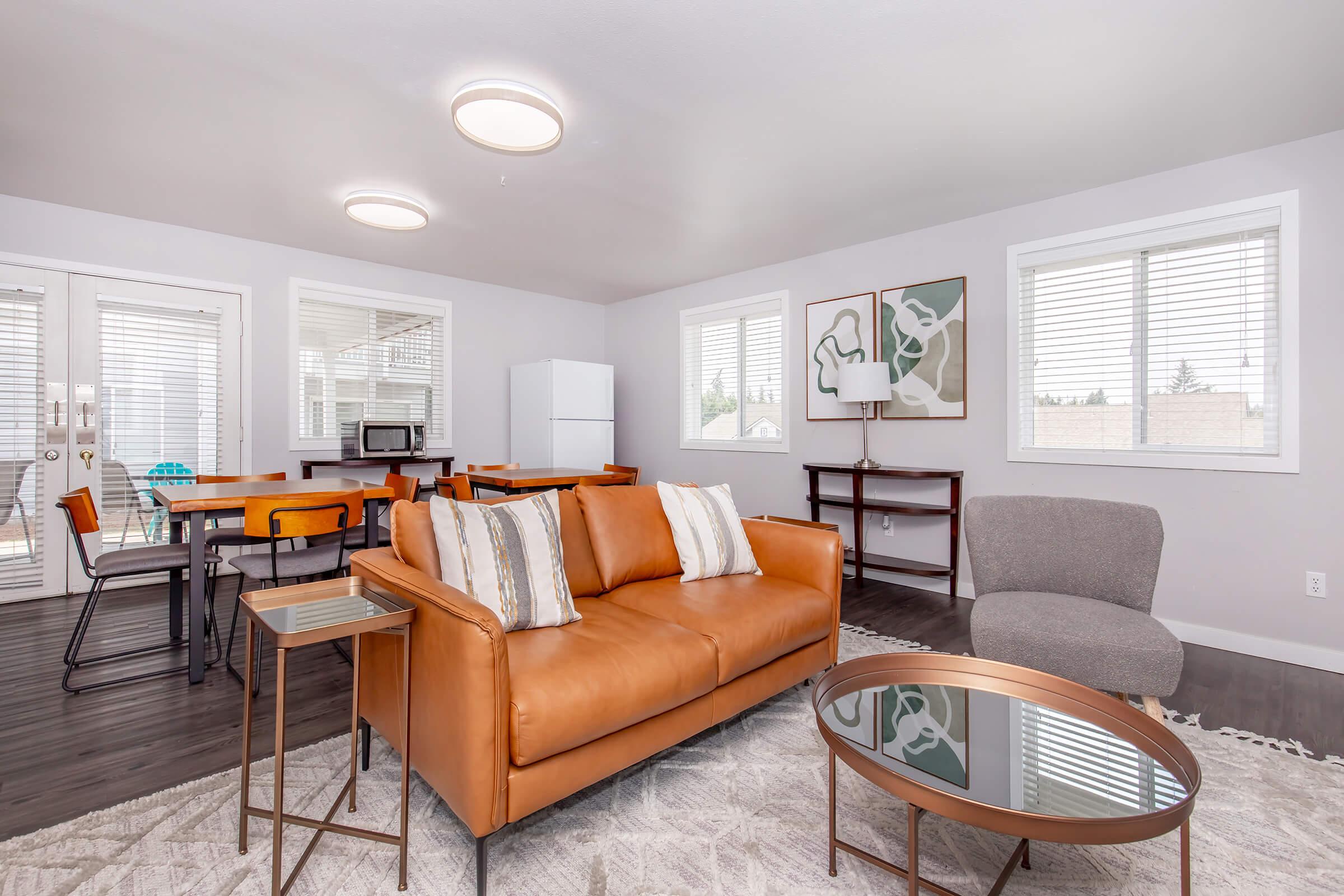 A modern living room featuring a brown leather sofa, a glass coffee table, and a gray armchair. In the background, there is a dining area with a table and chairs, a microwave, and a white refrigerator. Large windows allow natural light into the space, which has a minimalist design with neutral colors.