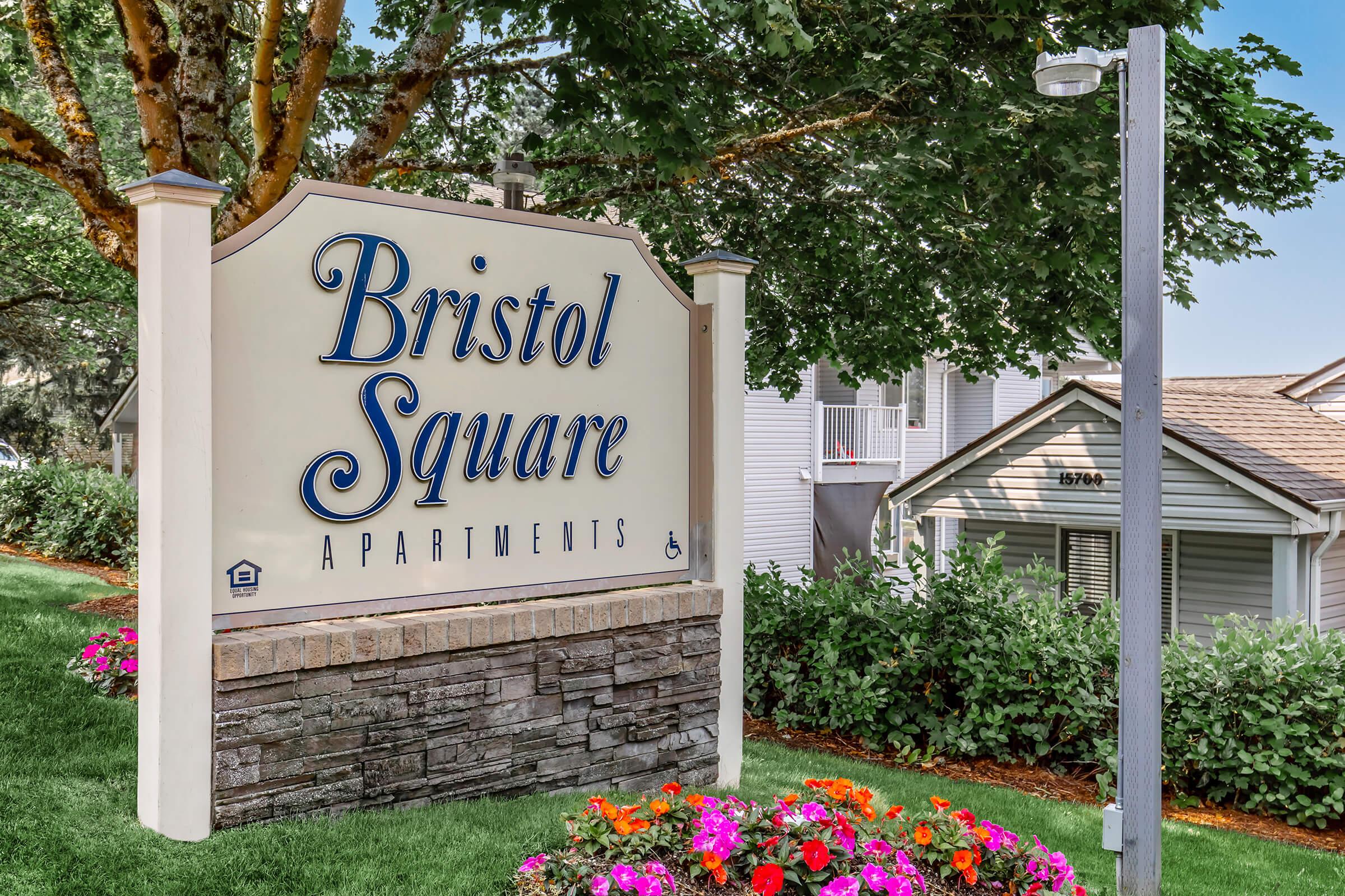 Sign for Bristol Square Apartments featuring the name in bold blue letters. The sign is set against a lush green backdrop with trees and colorful flowerbeds below. The surrounding area includes residential buildings, indicating a welcoming living environment.